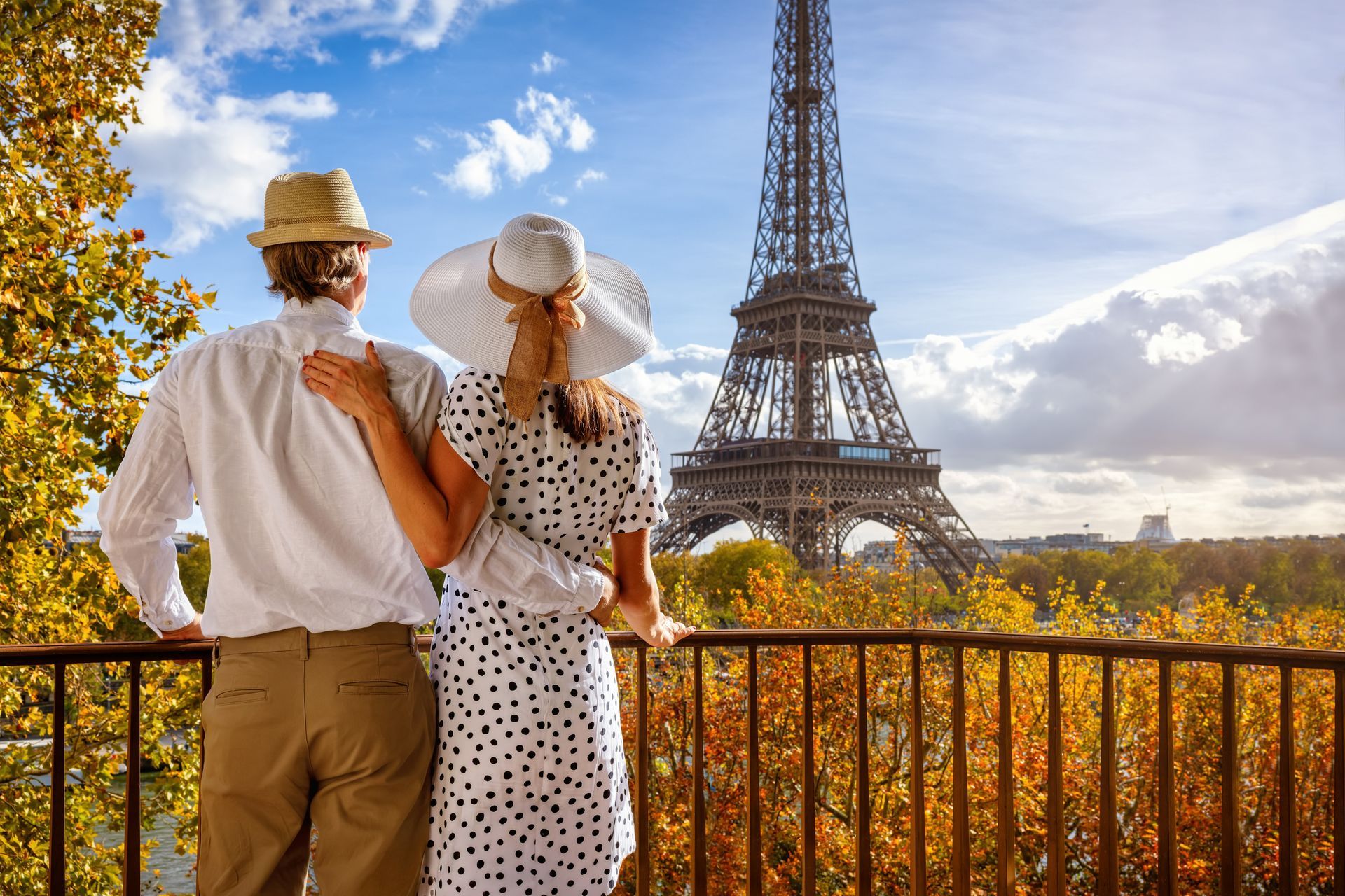 Couple in hats admiring the Eiffel Tower from a balcony with autumn trees below