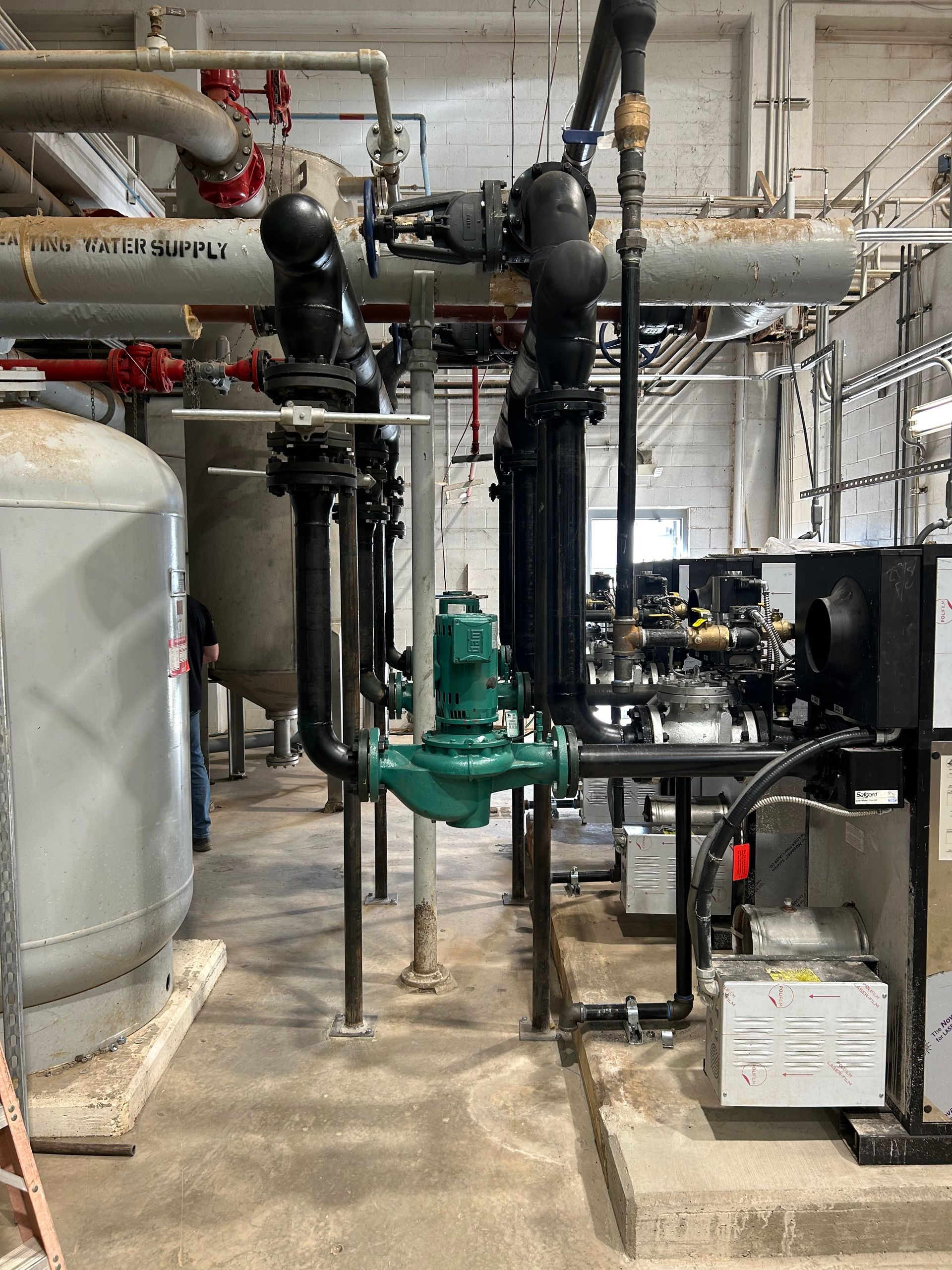 Industrial piping and equipment in a utility room. Green valve, black pipes, and silver tanks.