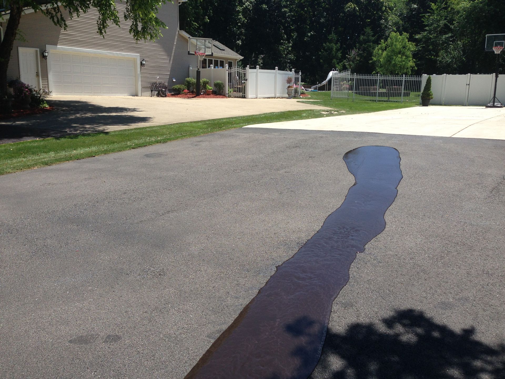 A driveway with a long puddle reflecting the sky and shadows; a house and basketball hoop are in the background.
