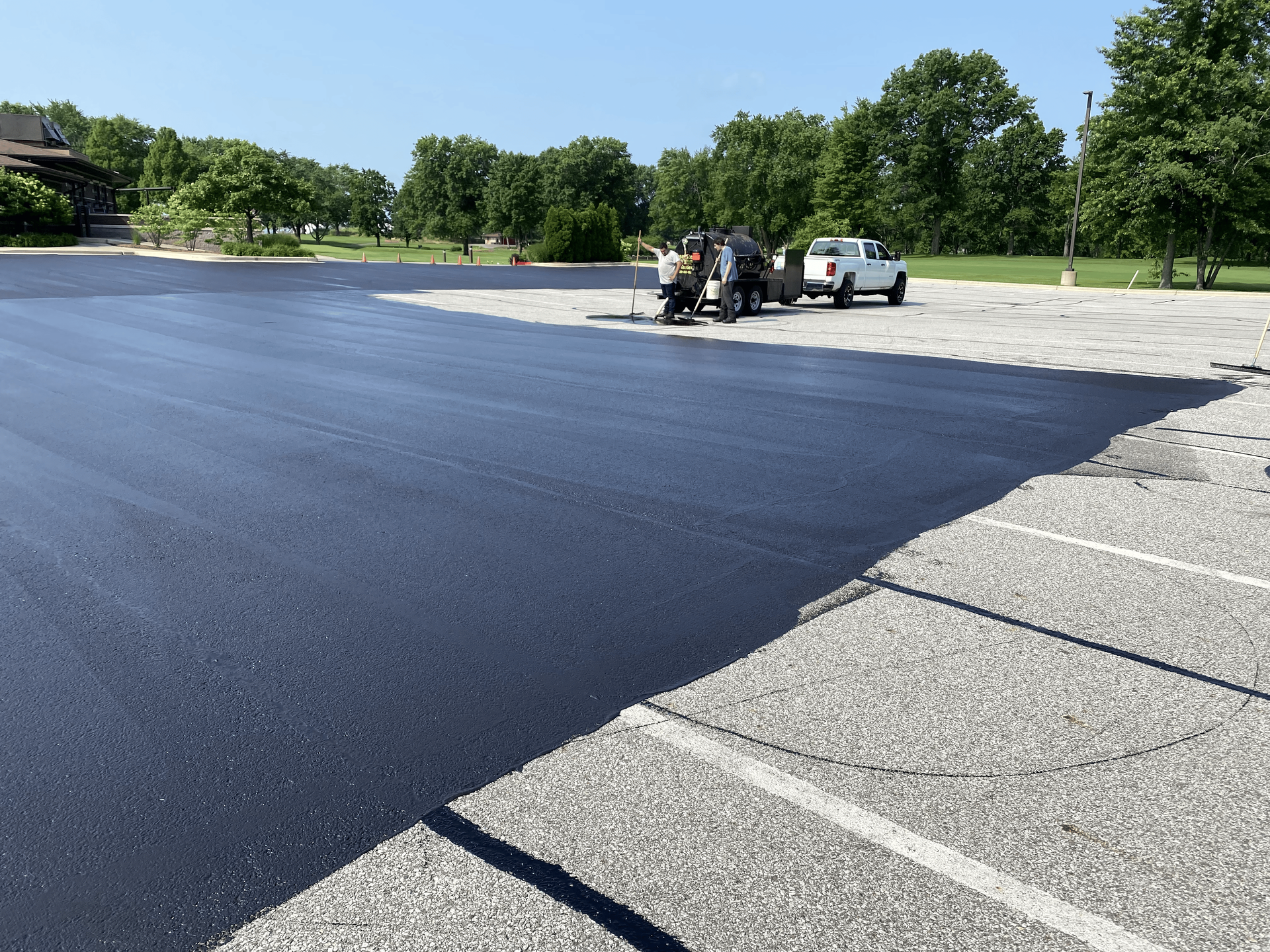 Freshly paved asphalt parking lot; workers and a truck visible; green trees in the background.