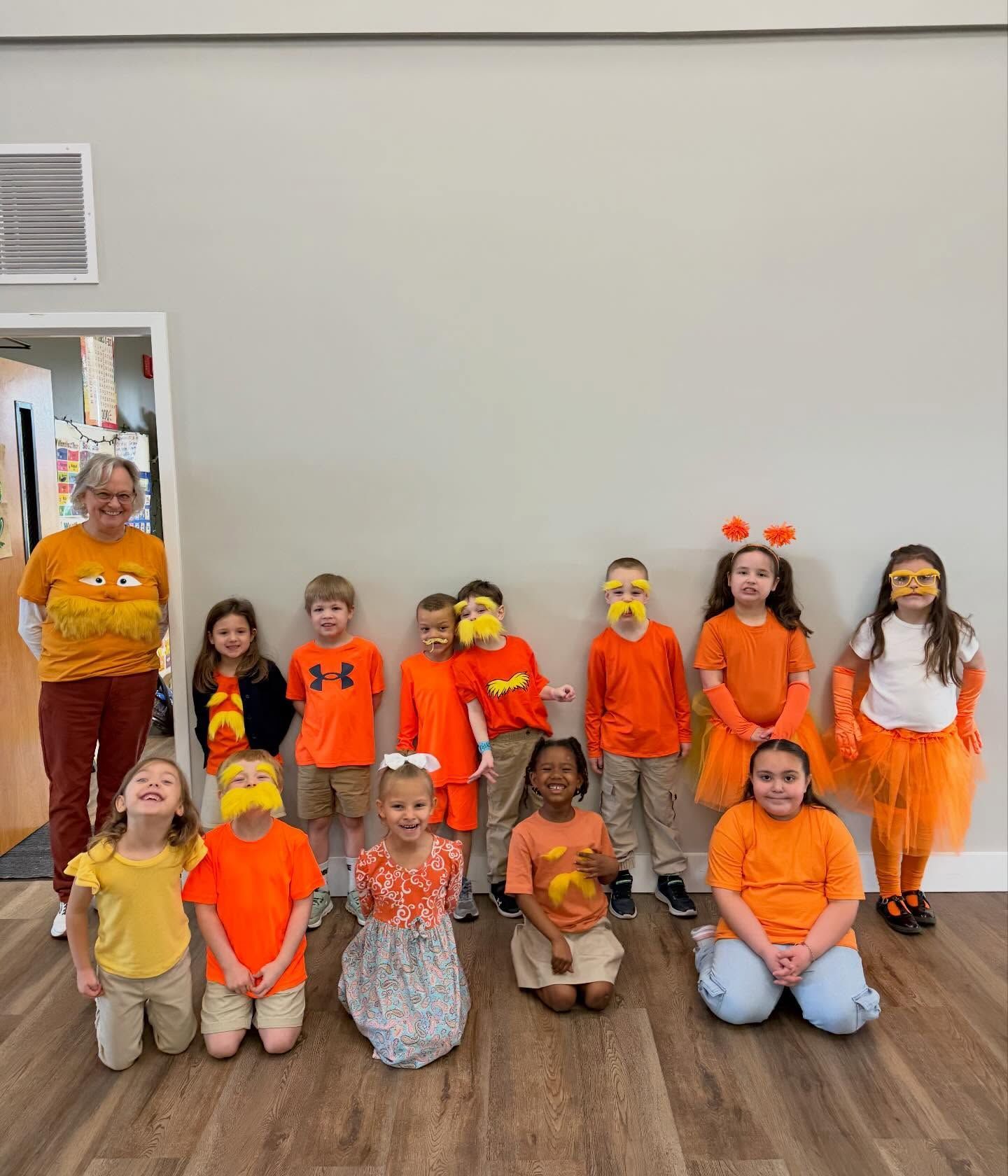 Children in orange costumes and a teacher posing in a room with a wood floor, some children kneeling in front.