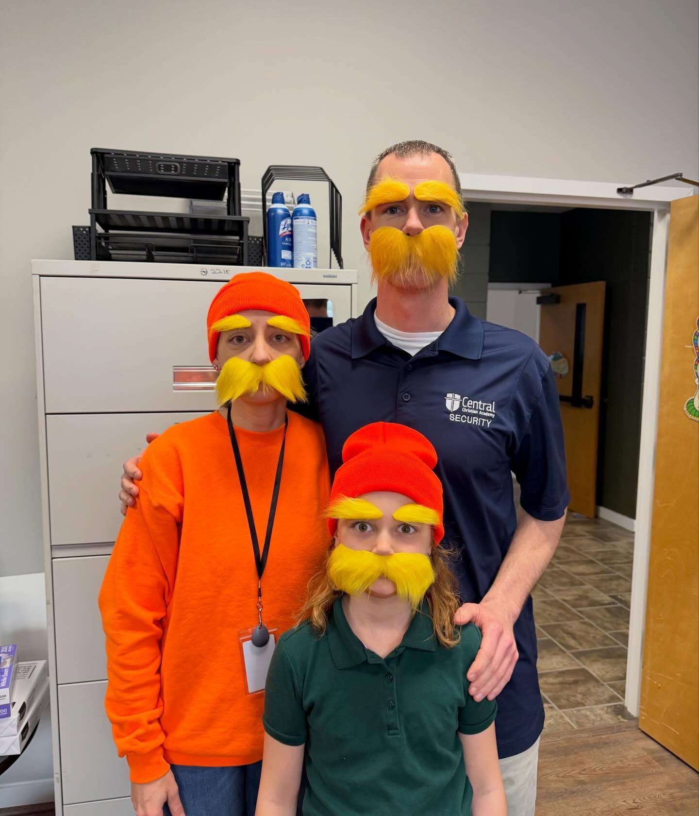 Three people in a room wearing red hats and yellow fake mustaches, posing together.