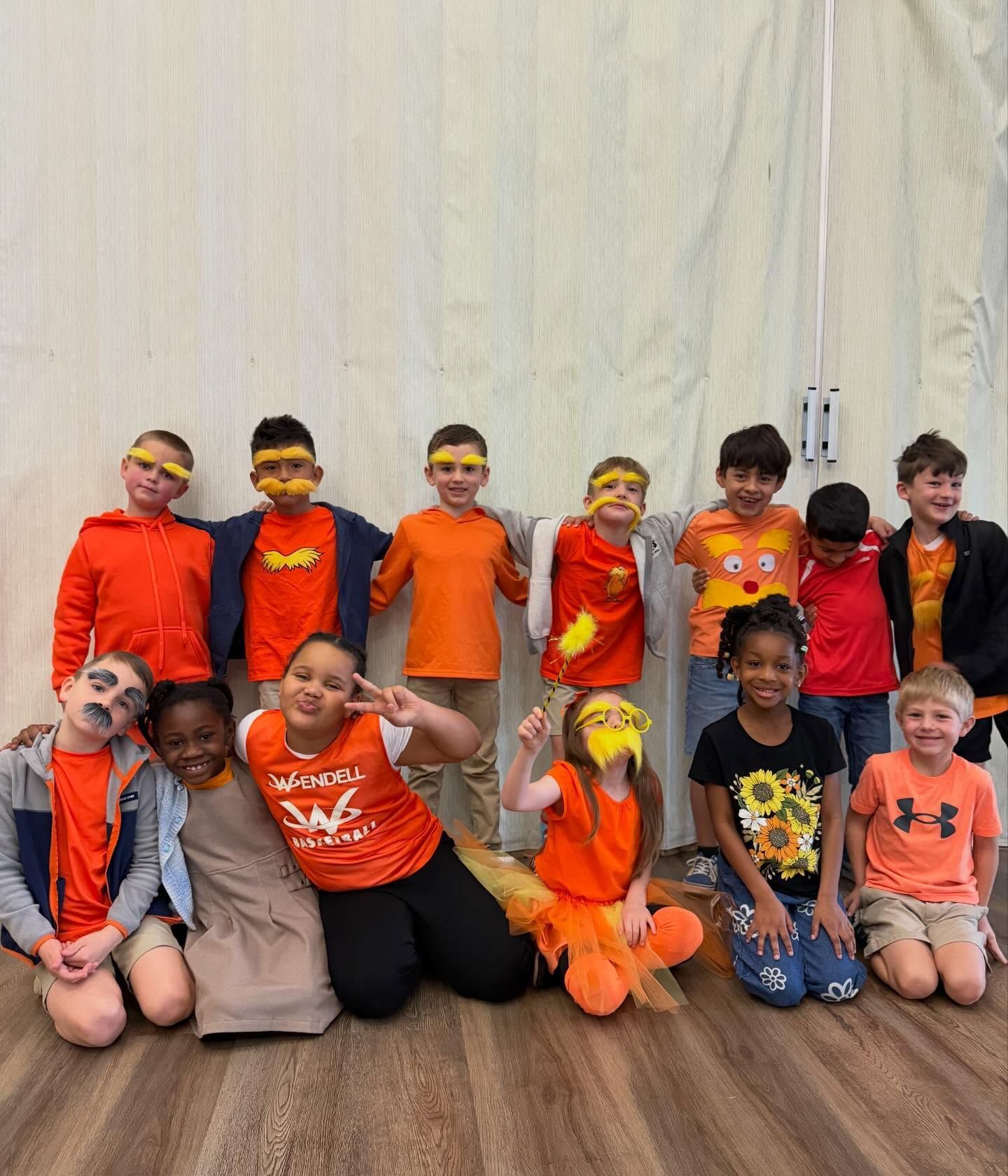 Group of children in bright orange costumes posing indoors against a light curtain backdrop.
