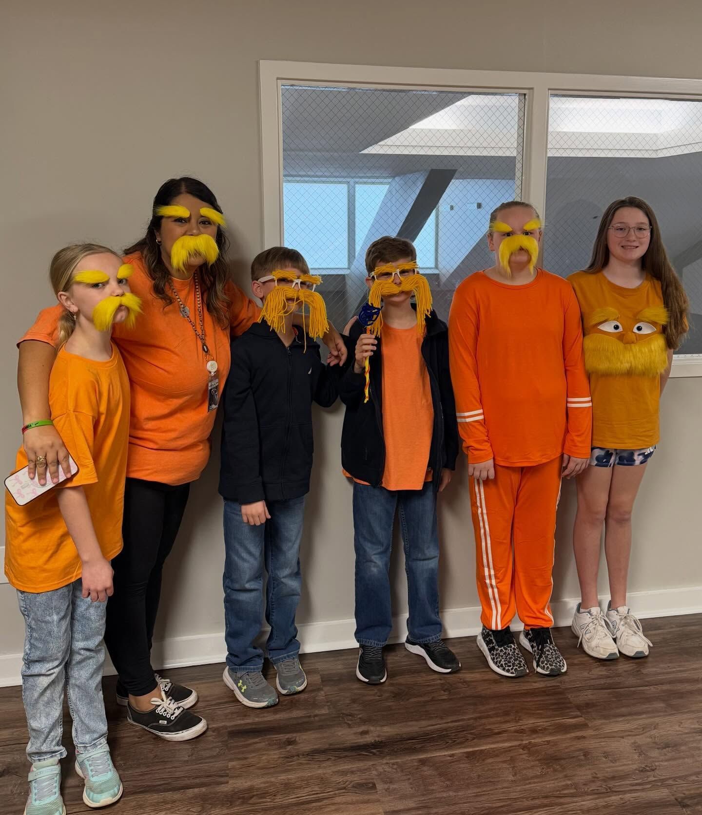 Seven people in orange and black shirts stand indoors, each wearing yellow goggles and holding orange snacks.