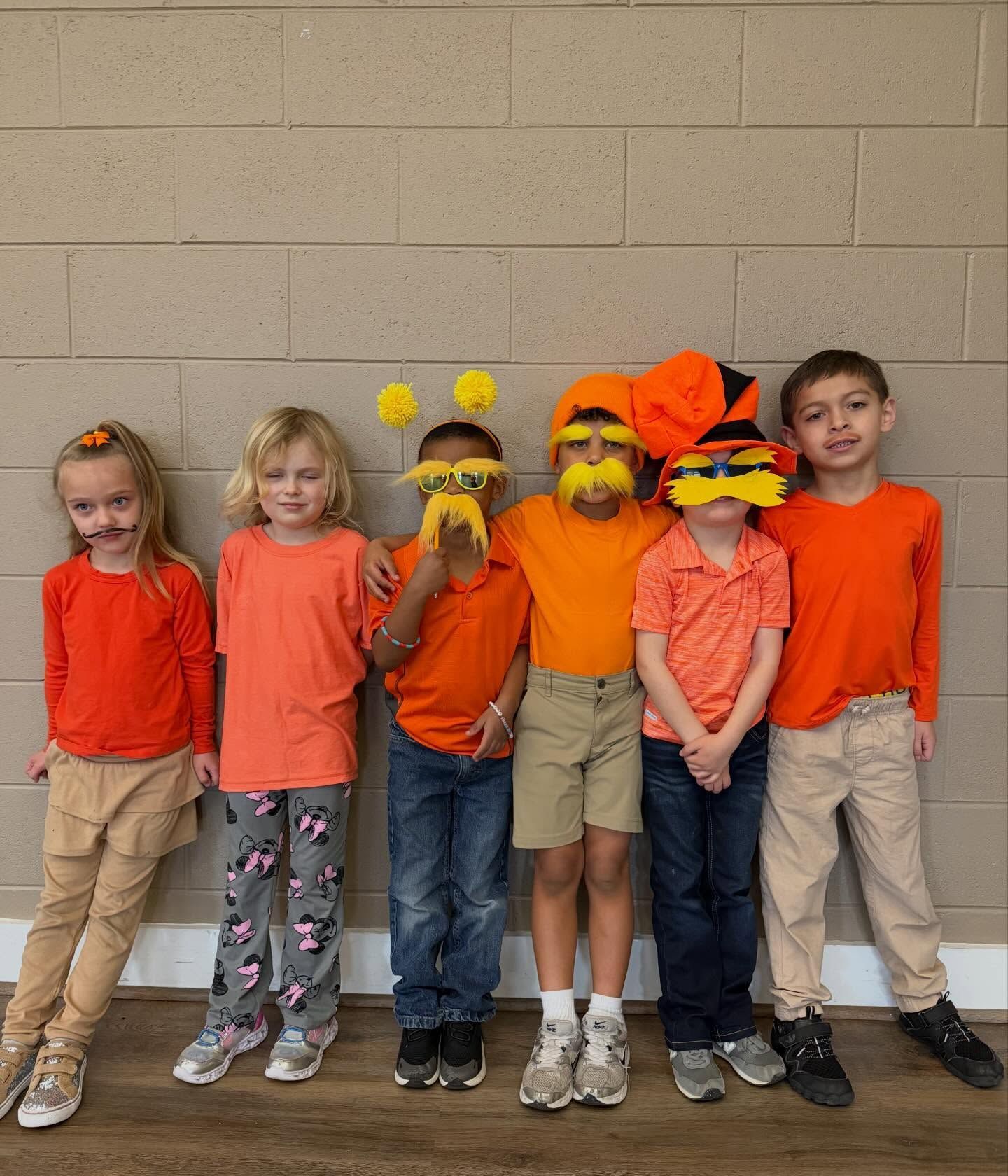 Five children in orange and tan outfits stand against a cinderblock wall, two wearing colorful bee-like costumes.
