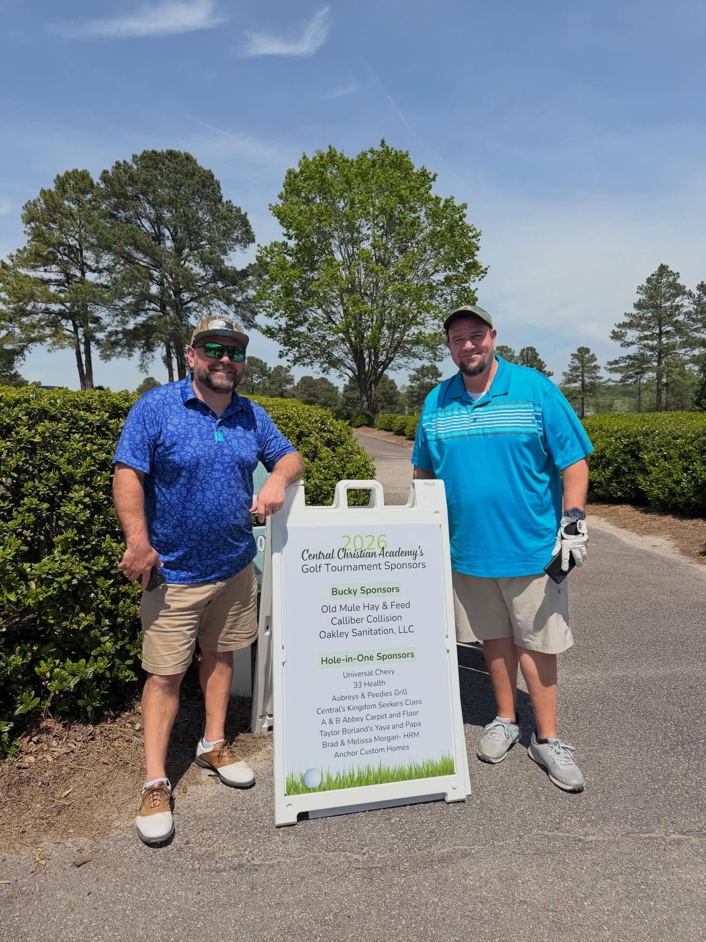 Two men standing beside a roadside sign on a sunny golf course.