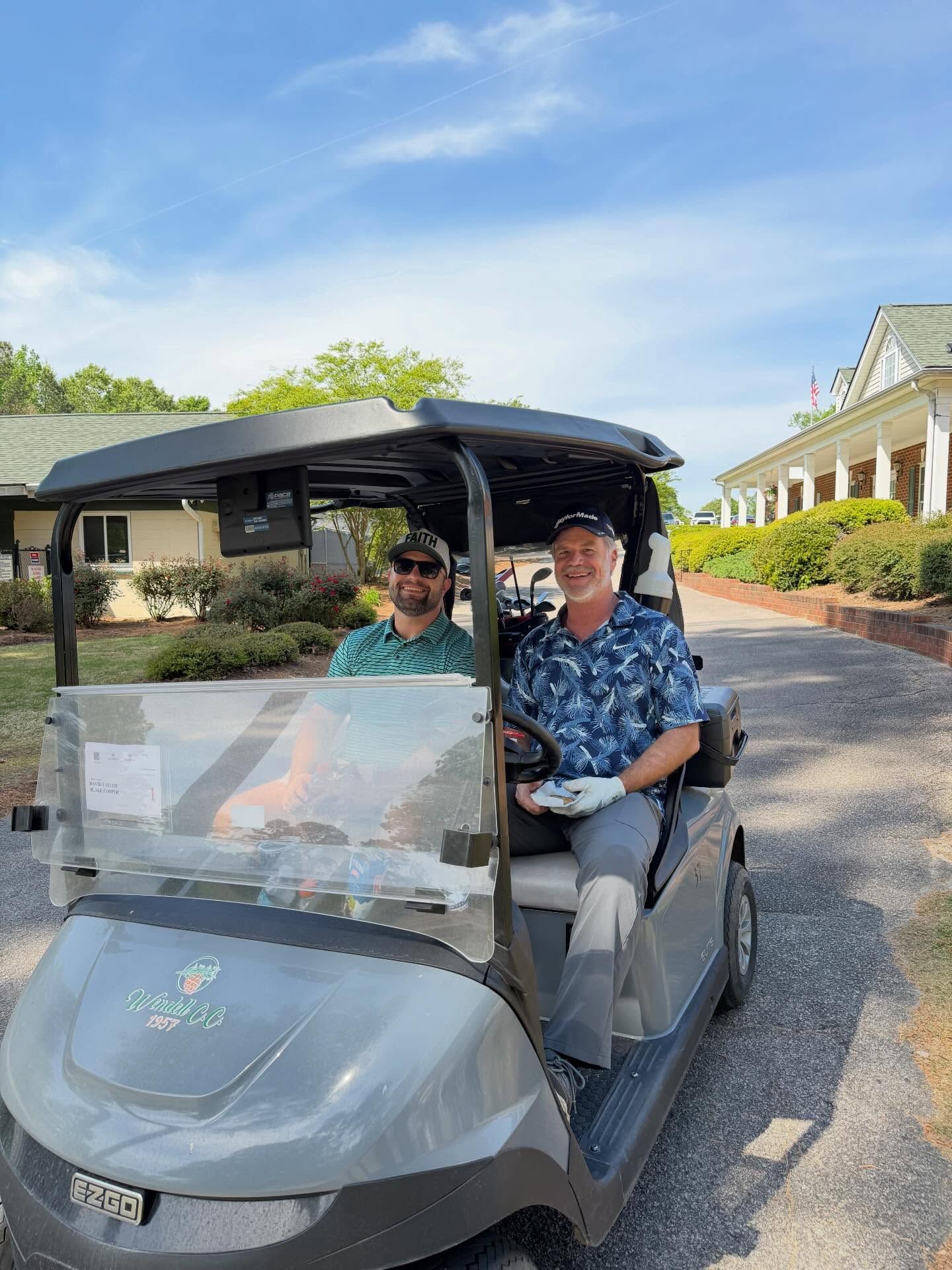 Two people riding in a gray golf cart on a sunny driveway beside a building