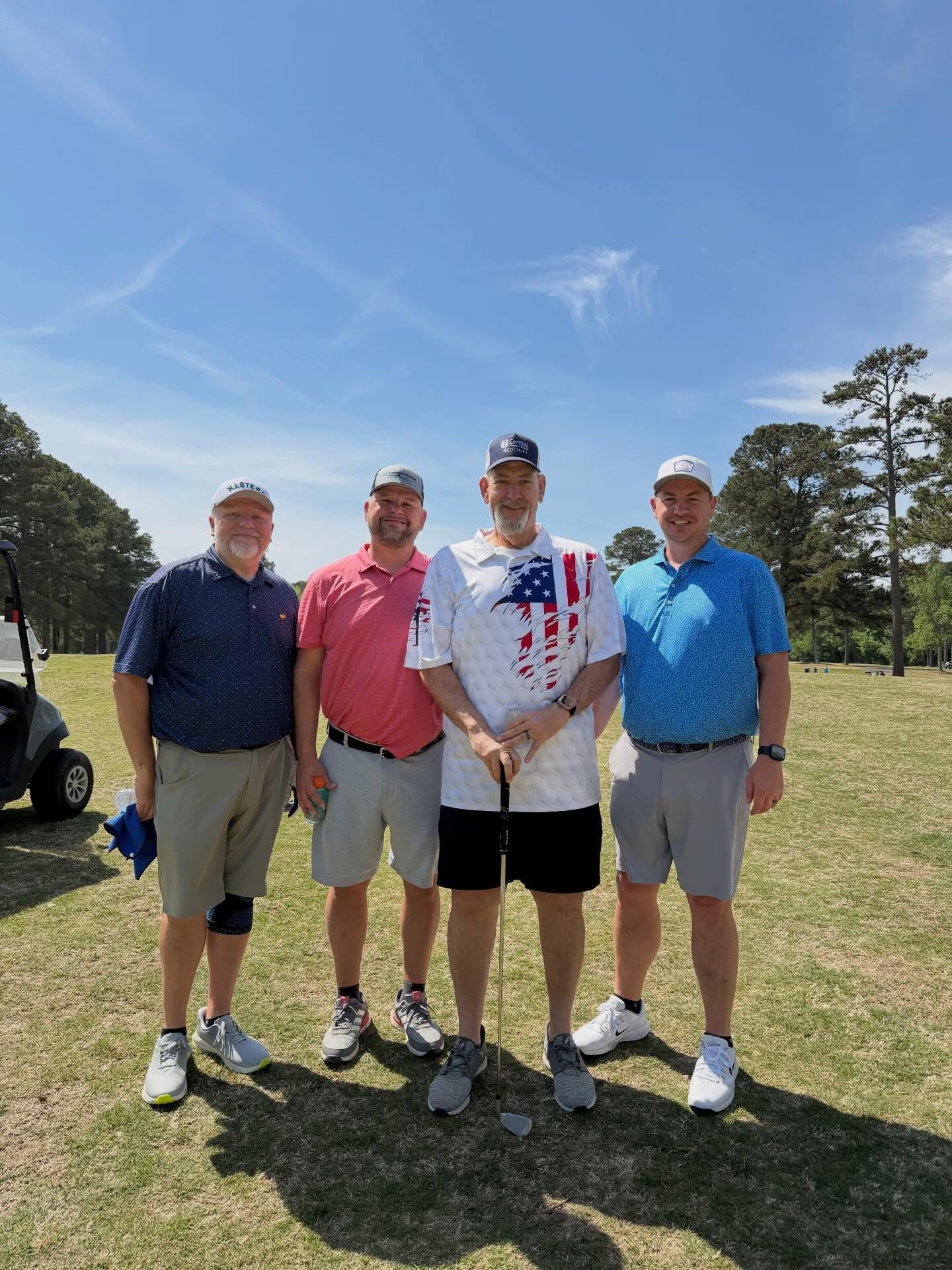 Four men posing on a sunny golf course, wearing casual shirts and shorts under a blue sky.