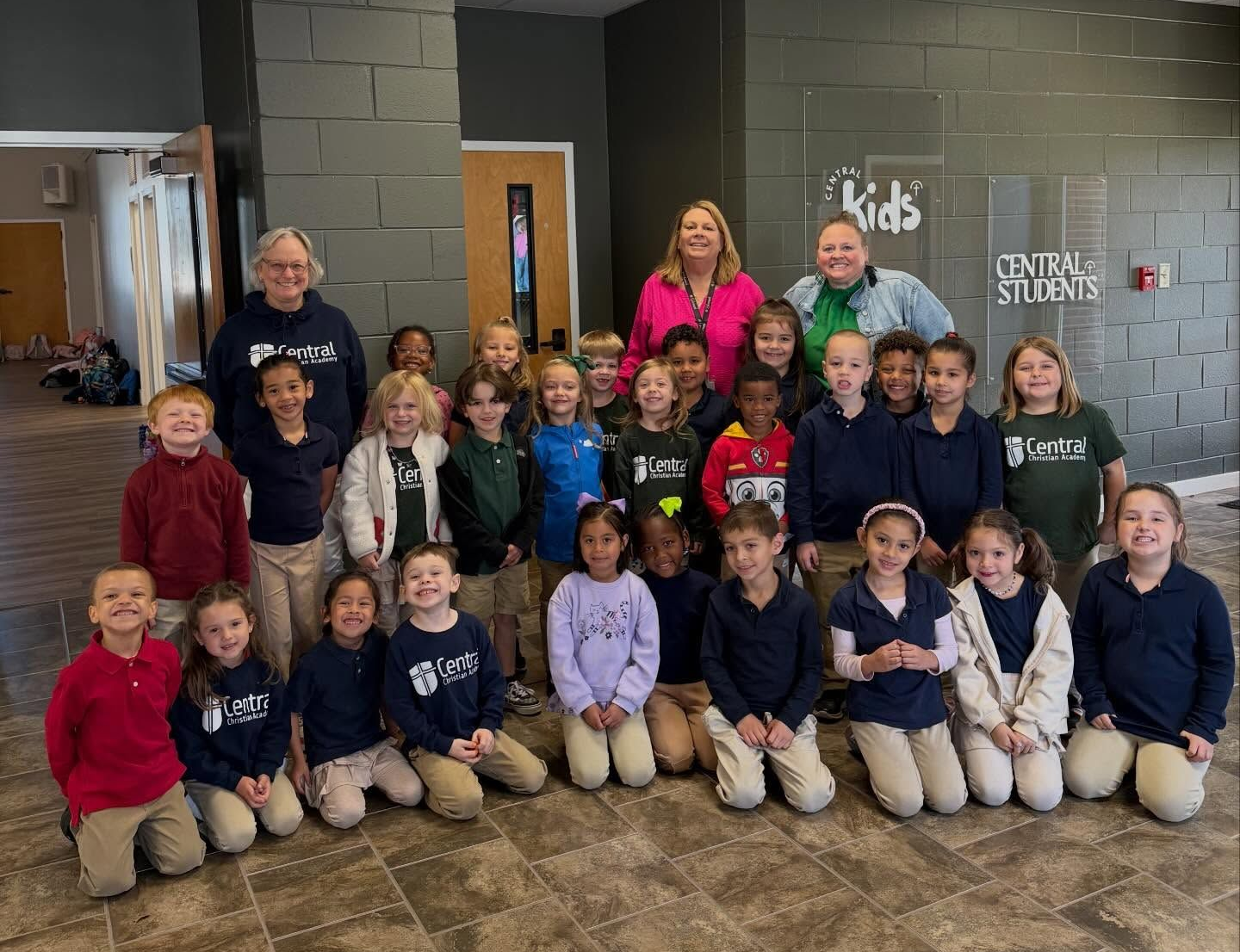 Group of schoolchildren and teachers posing in a hallway with gray brick walls and school signs.