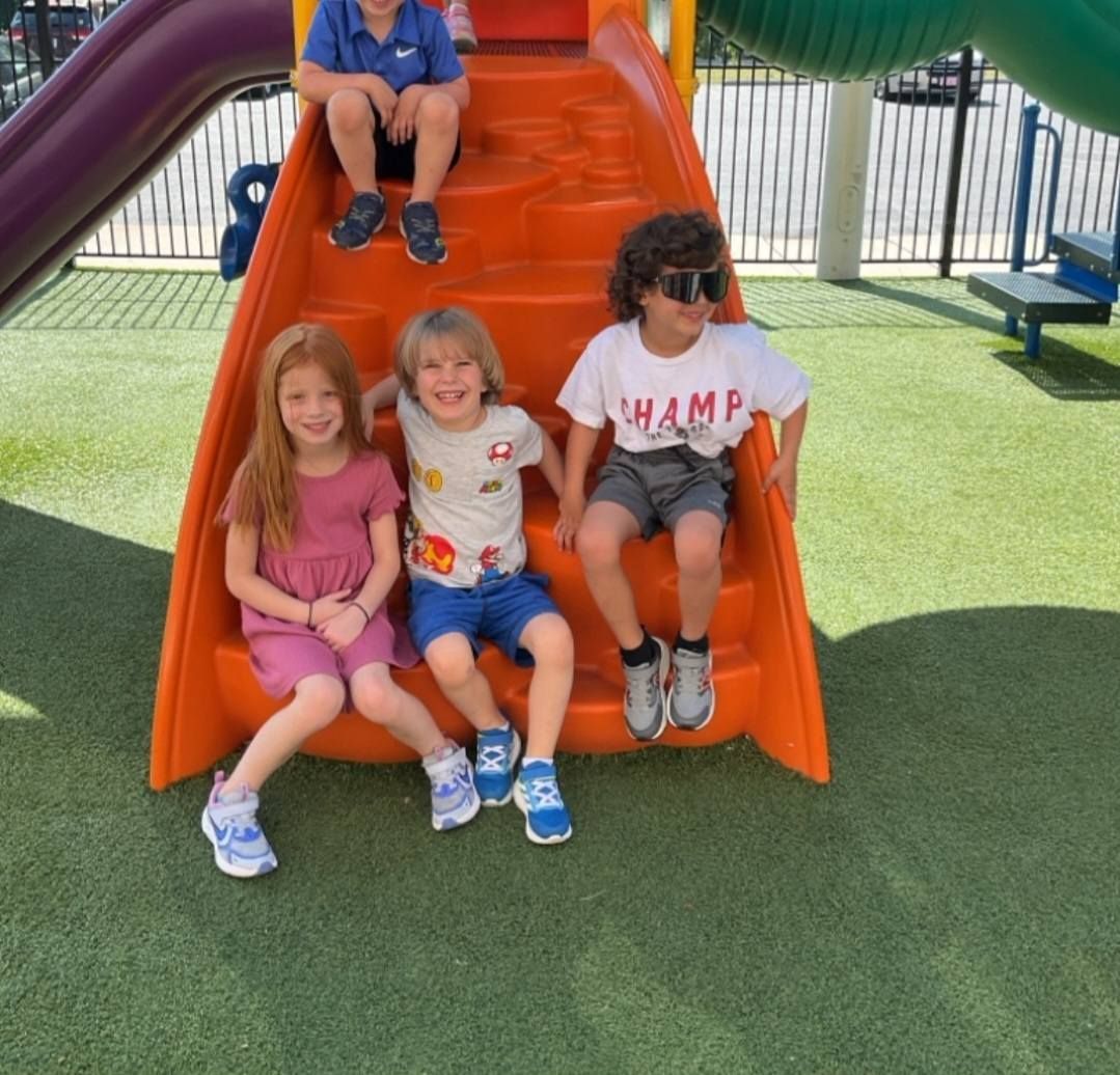 Children sitting on an orange slide at a playground, with green turf and a play structure behind them.