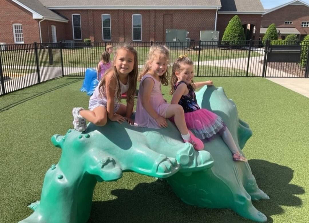 Three children sitting on a green turtle playground sculpture outdoors, smiling.