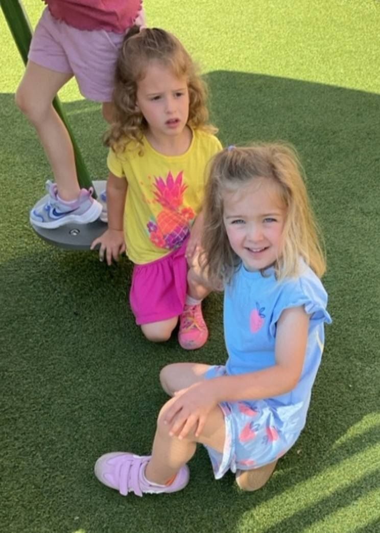 Two children crouch on green turf, smiling, with a third child’s legs visible behind them.