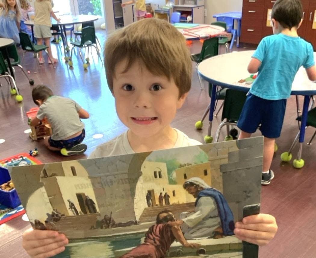 Child in a classroom holds a painting of a desert scene with figures and buildings.