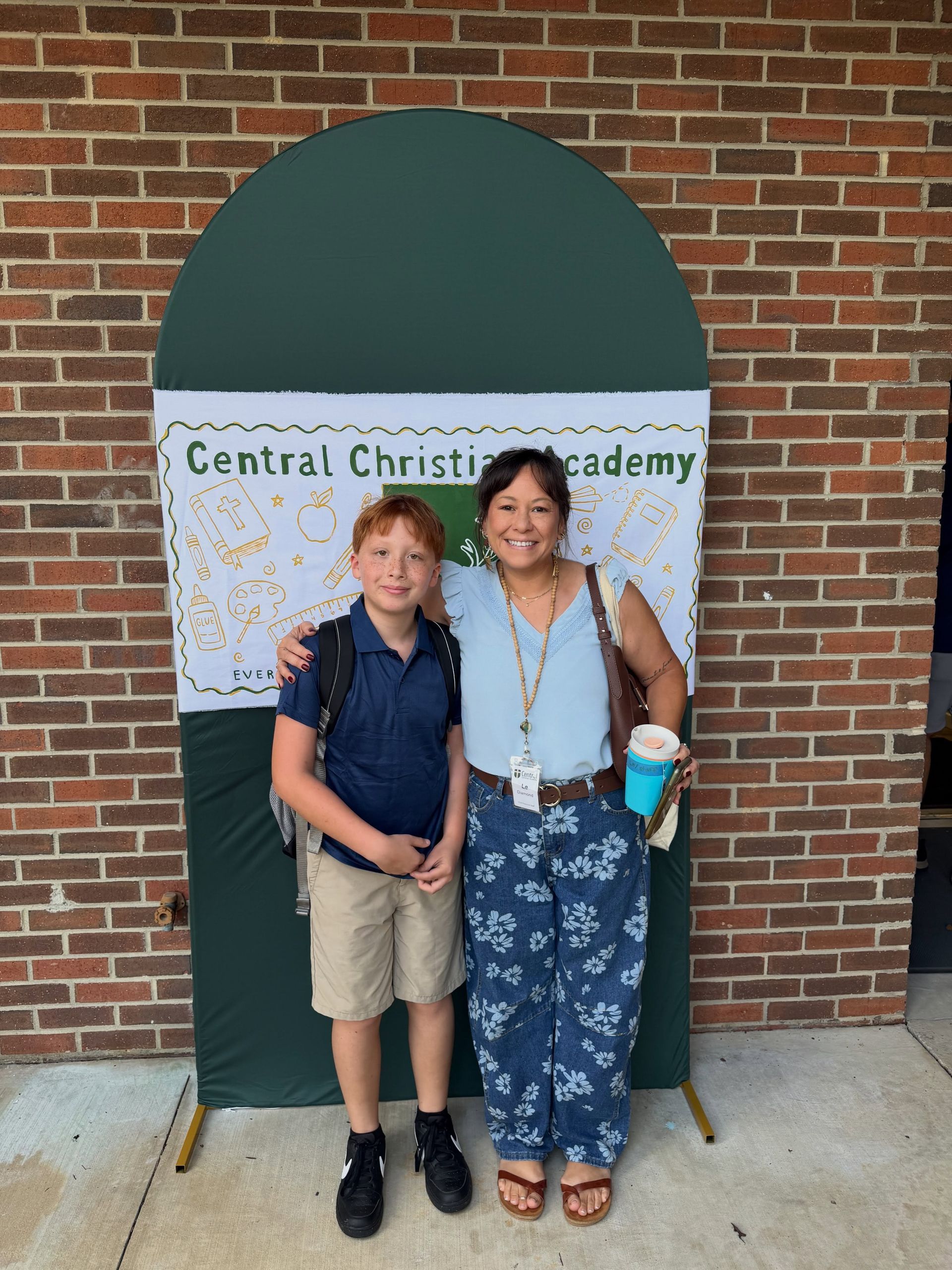 Two people posing in front of a Central Christian Academy sign, smiling outdoors by a brick wall.