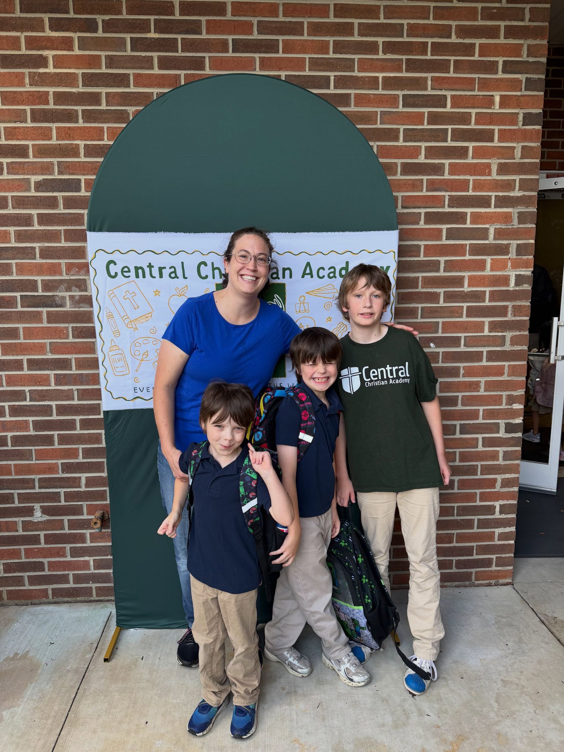 Four children standing in front of a brick wall and school sign, smiling at the camera.