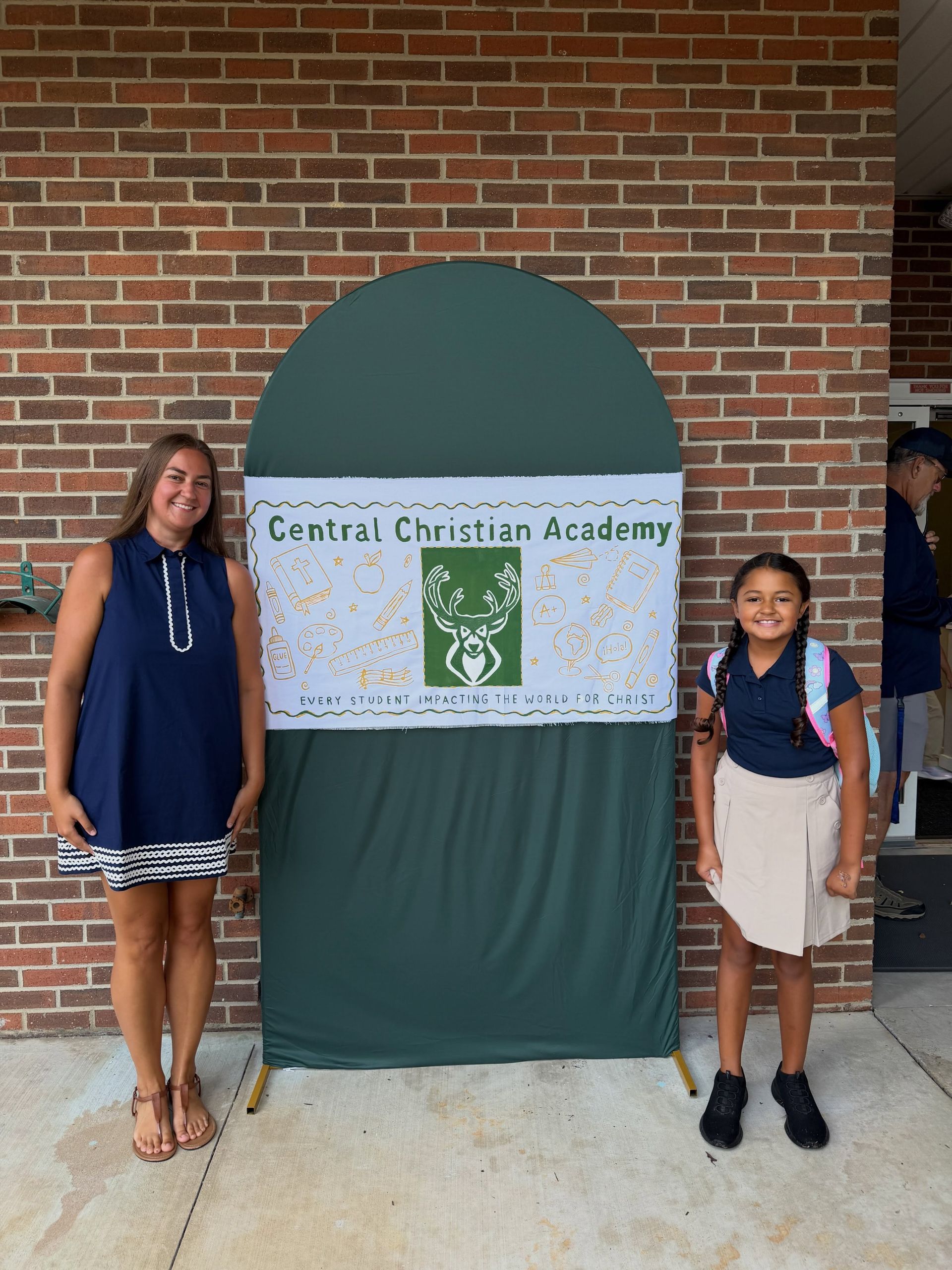 Two students standing beside a Central Christian Academy sign on a brick wall backdrop