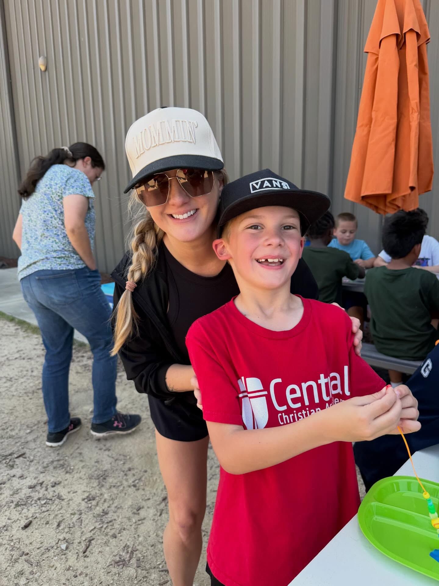 Woman and boy smiling outdoors at a community event, standing by a table with a green bowl