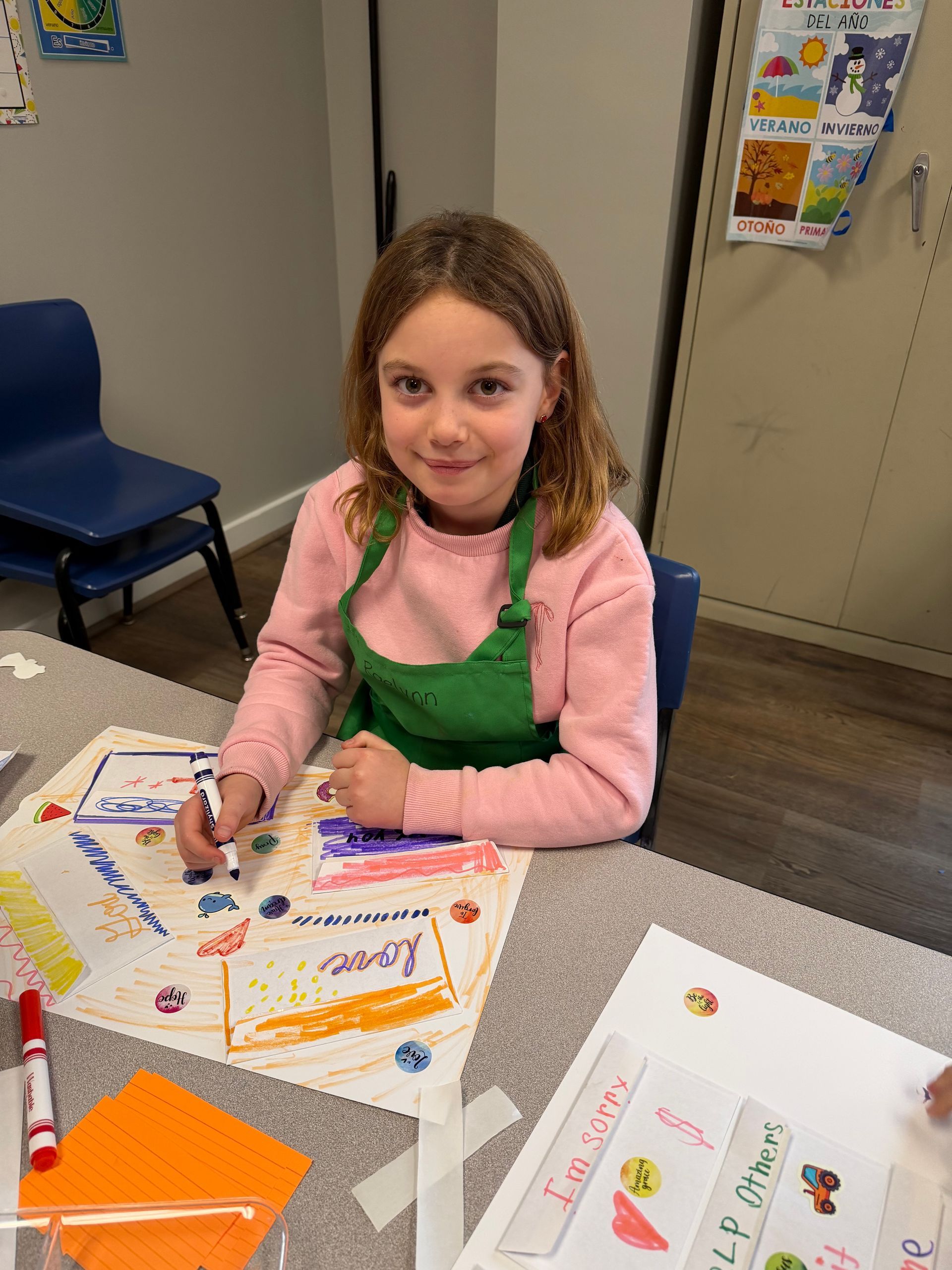 Child smiling at a table, coloring with markers and crayons in a classroom.