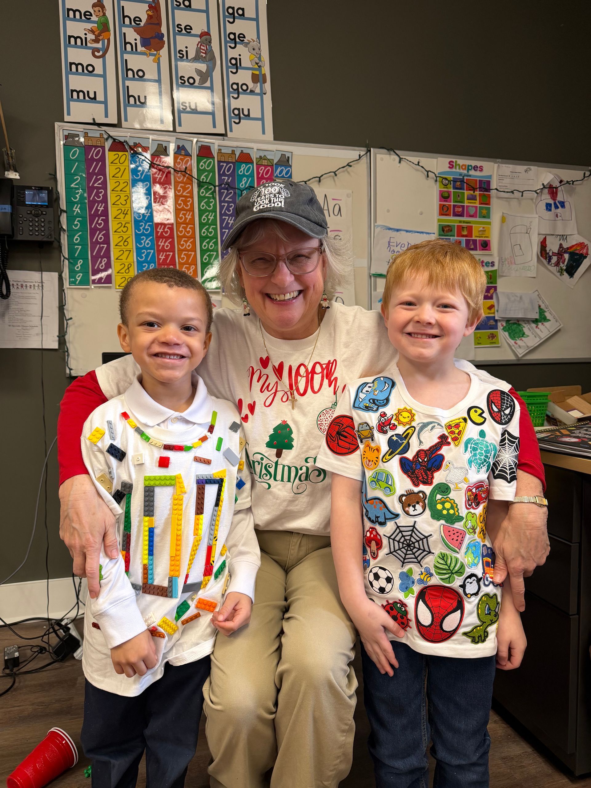 Woman seated with two smiling children in a classroom, colorful posters and bulletin boards behind them