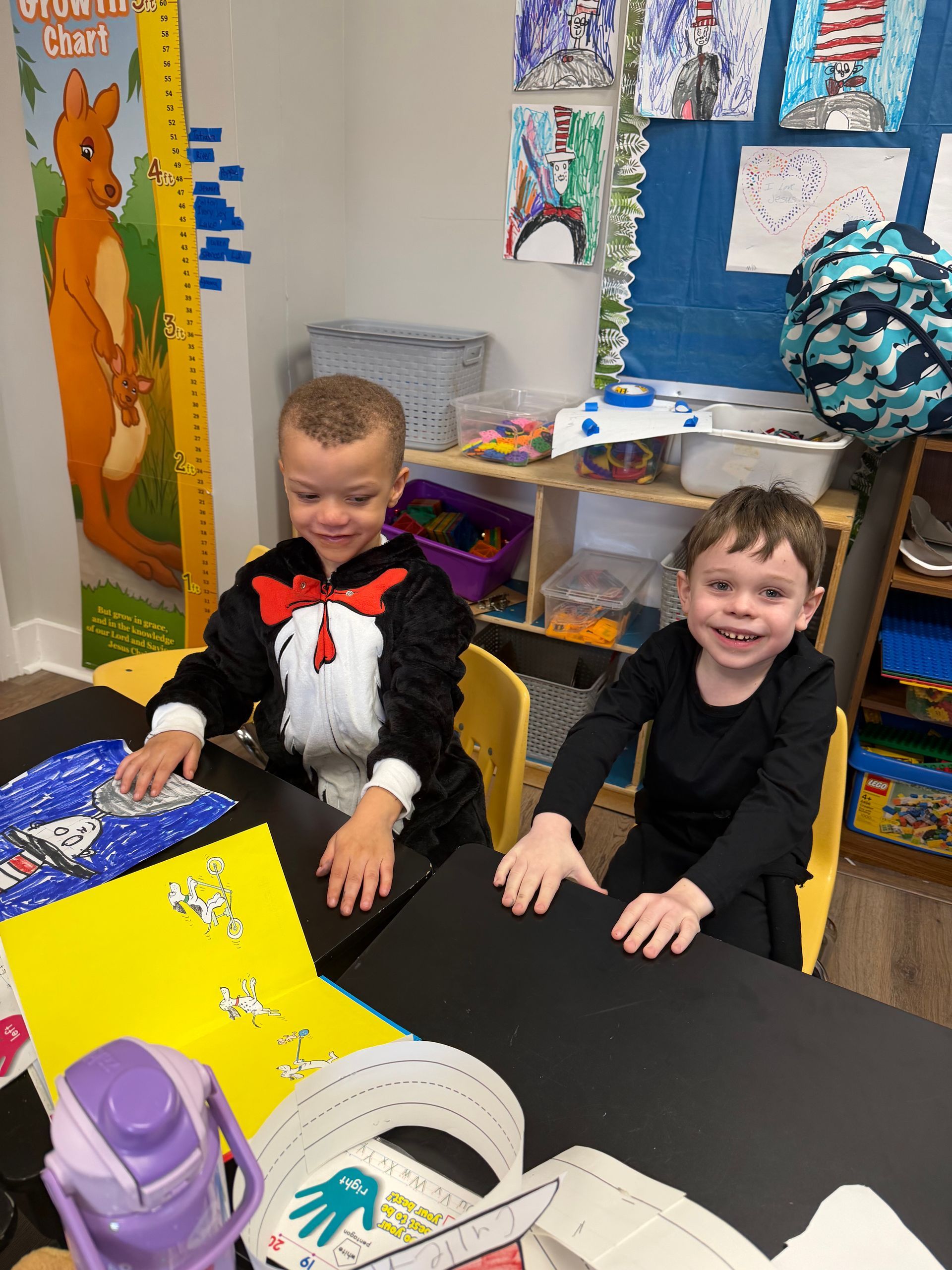 Two children smiling at a classroom table with craft supplies and colorful decorations.