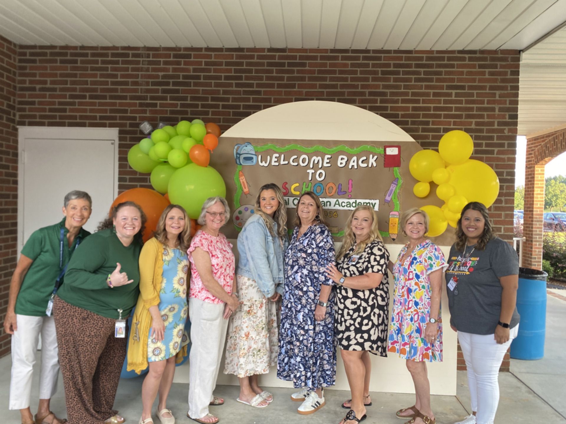 Group of people posing under a “Welcome Back” balloon banner at a brick building entrance