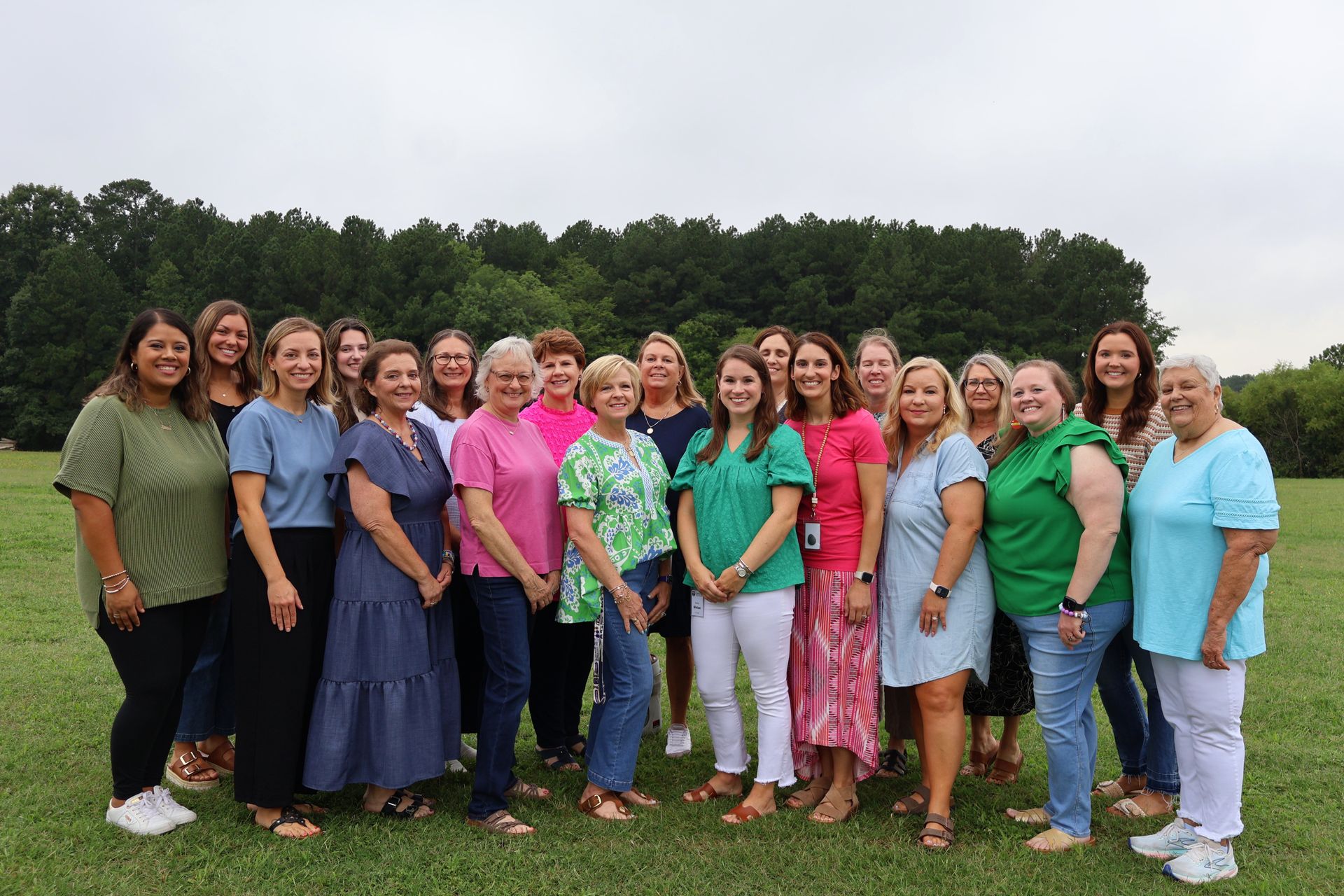 Group of people standing outdoors on grass, smiling in colorful shirts in front of trees.