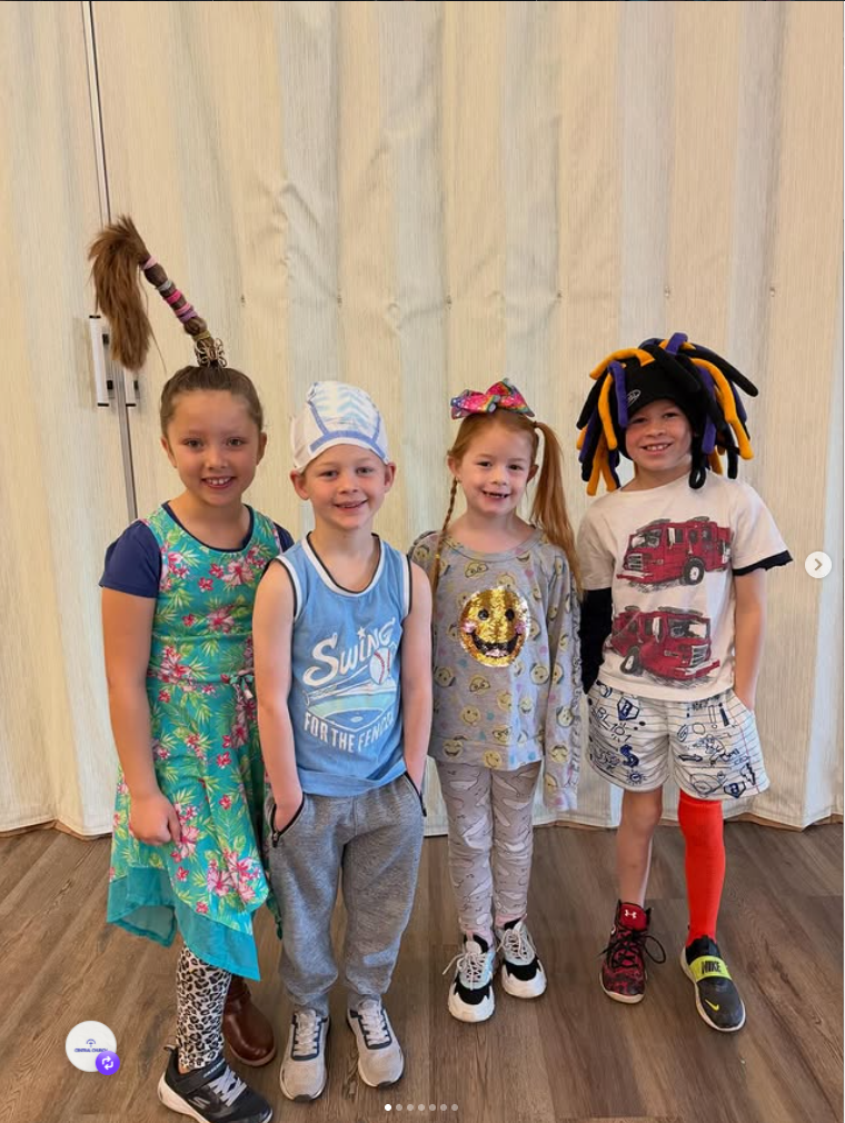 Four children stand smiling indoors in front of cream curtains, wearing colorful costumes and hats.
