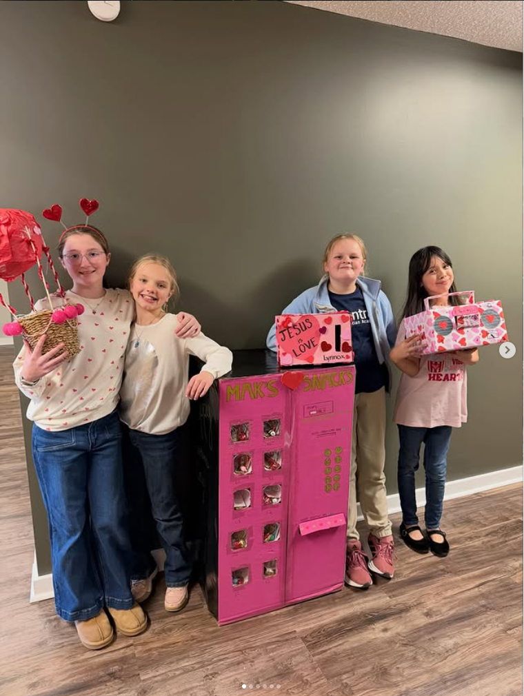 Four girls pose indoors beside a bright pink display stand with Valentine’s-themed gift boxes.