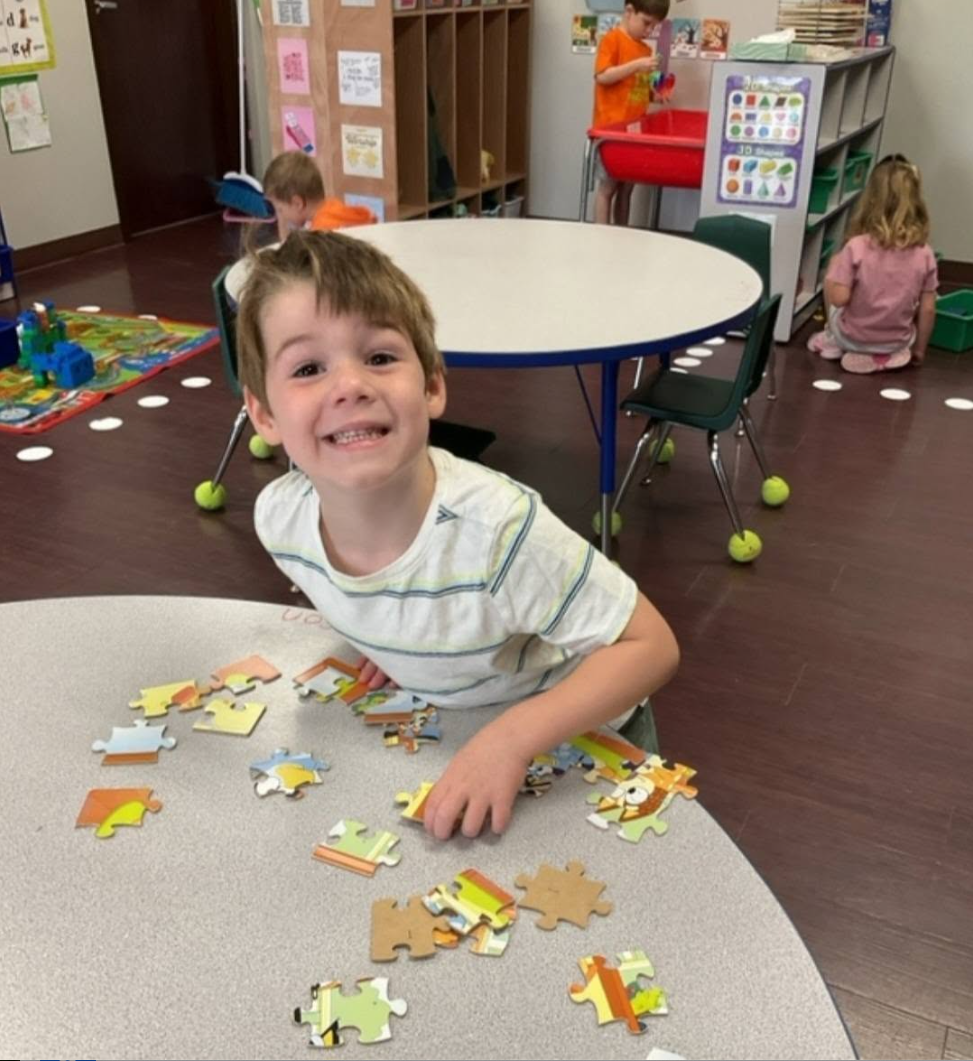 Child smiling at a table, coloring with markers and crayons in a classroom.