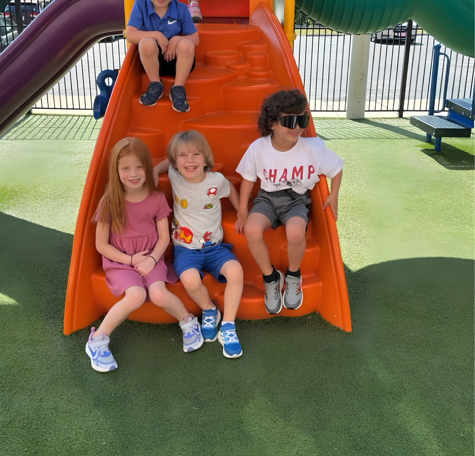 Three children sitting at the bottom of an orange playground slide, smiling outdoors.