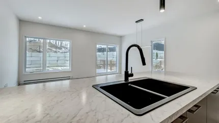 Cuisine moderne avec évier noir et robinet sur un comptoir blanc, donnant sur les fenêtres.