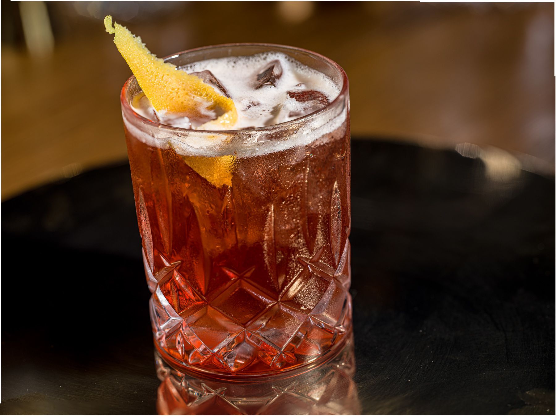 A glass of red liquid with ice and an orange peel on a table.