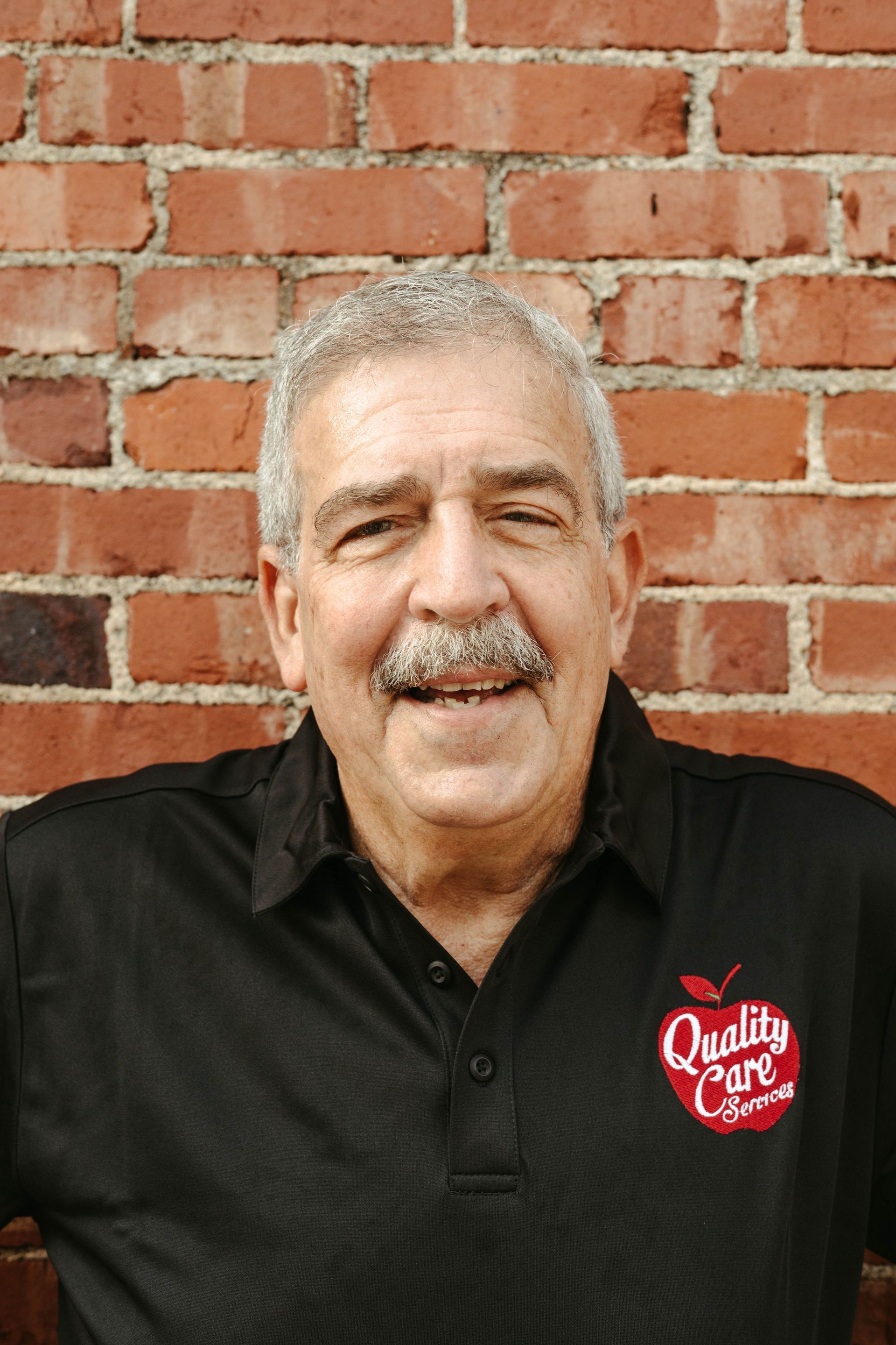 A man with a mustache is smiling in front of a brick wall.
