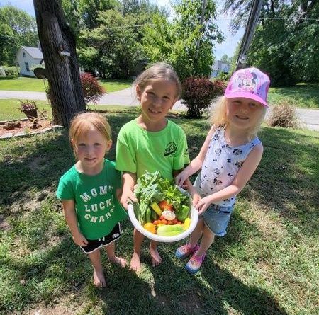 Three young girls are standing in the grass holding a bowl of vegetables.