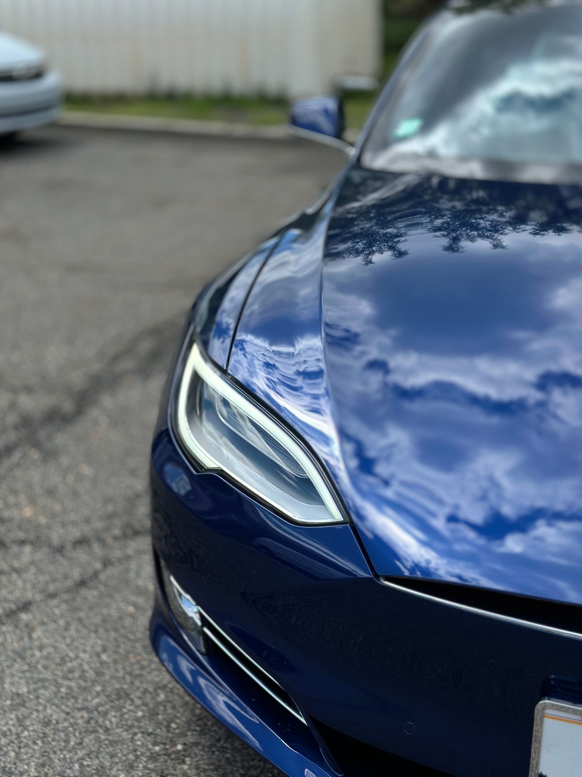 Close-up of a blue car’s front headlight and hood parked on pavement.