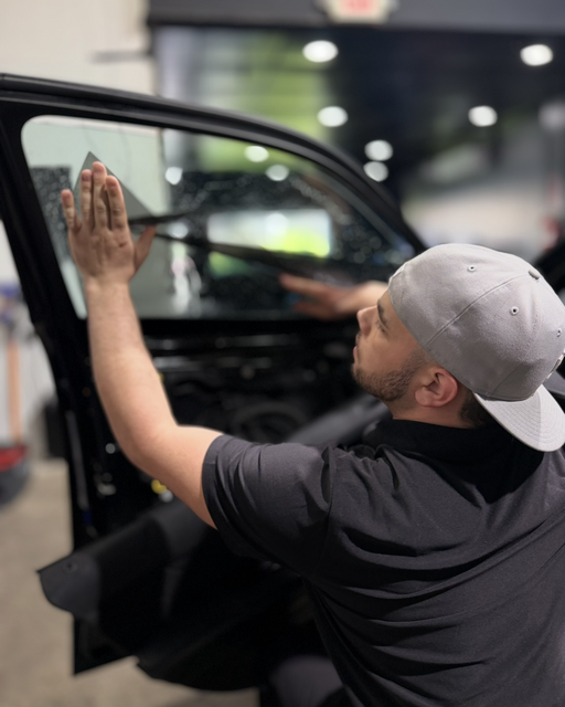 Man cleaning a car window with a cloth inside a garage, wearing a gray cap and black shirt