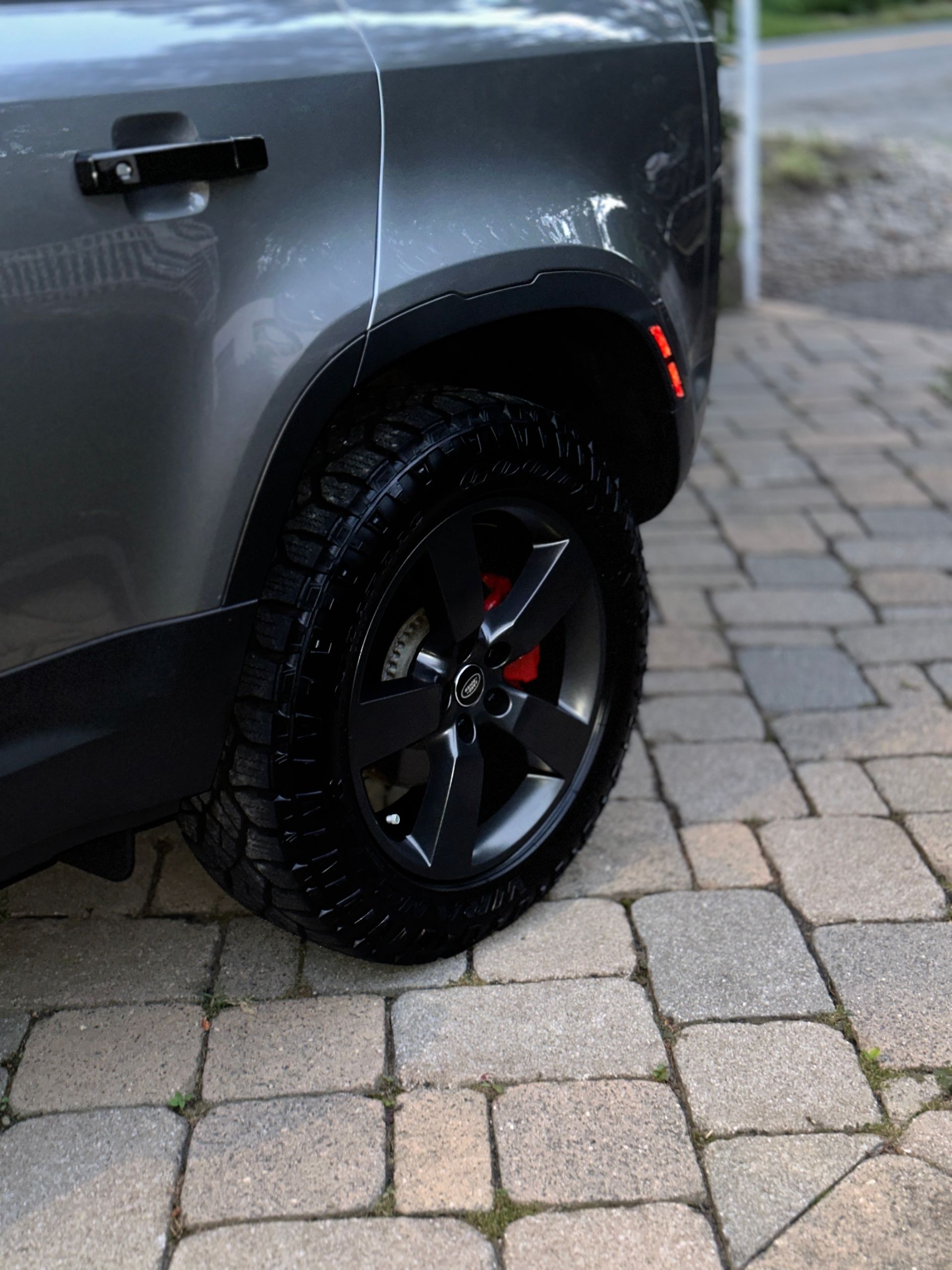 Close-up of a gray SUV’s rear wheel and fender on a cobblestone driveway.