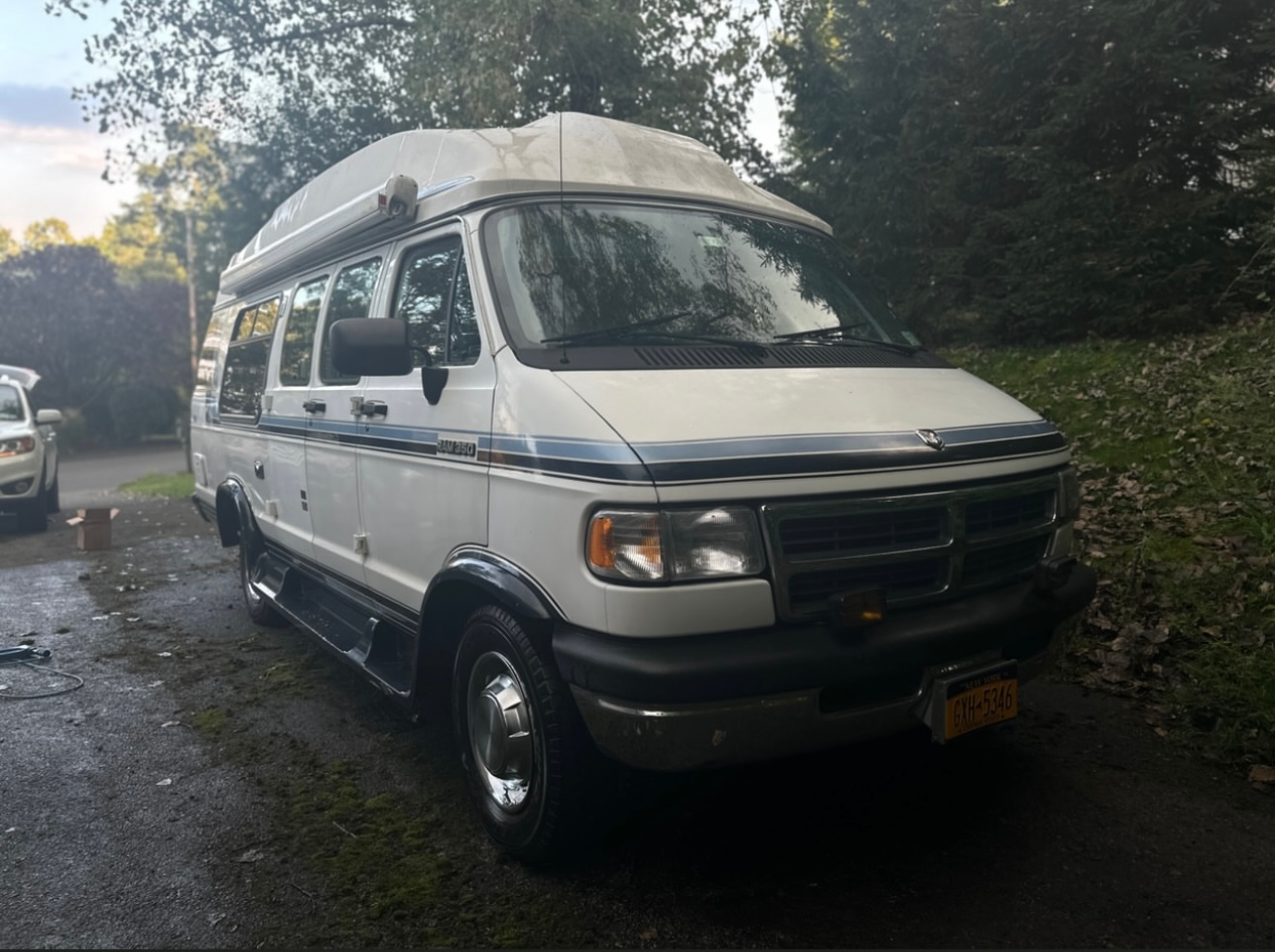 White passenger van parked on a roadside under a blue sky, with a car and orange cone ahead.