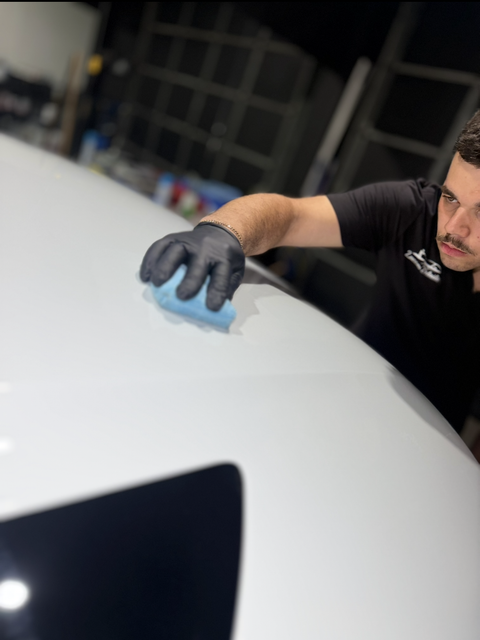 A man in black gloves wipes a white car surface in a garage.