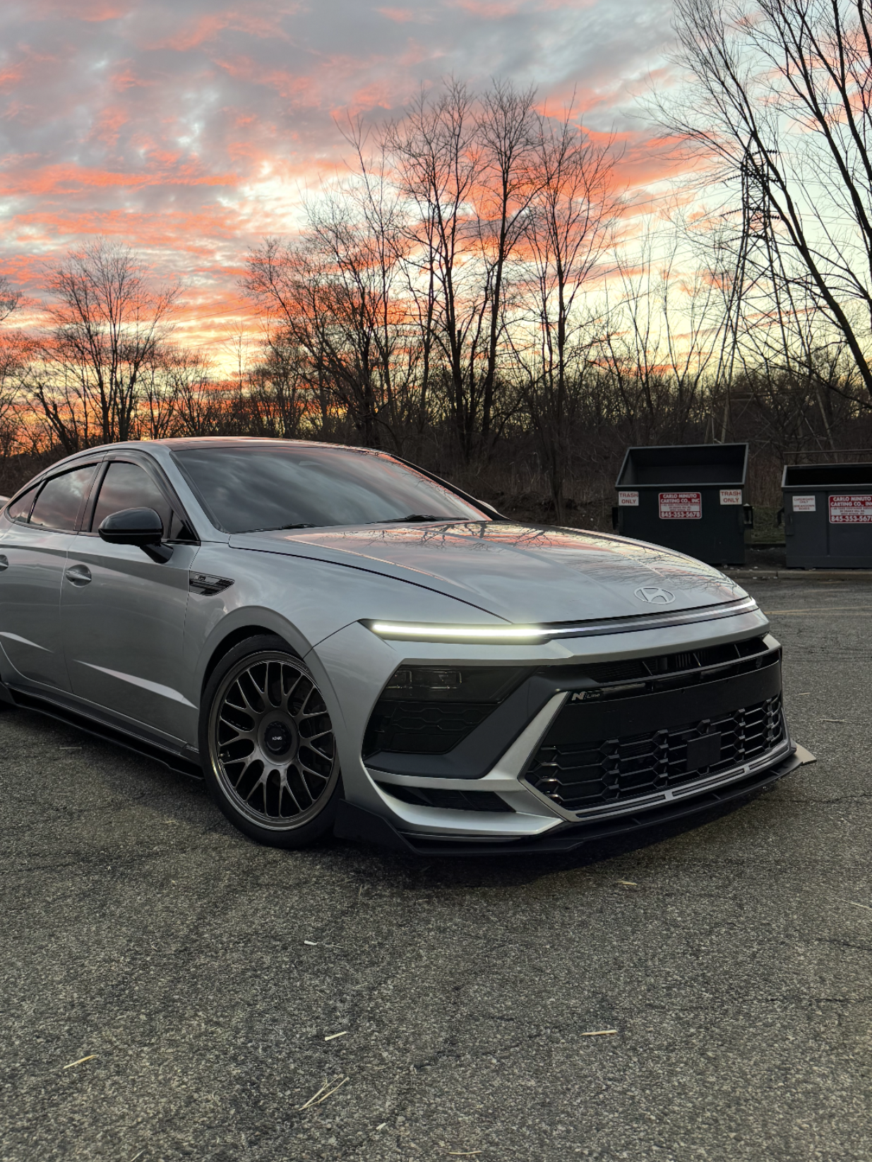 Silver sports car parked in a lot at sunset with pink clouds and bare trees in the background