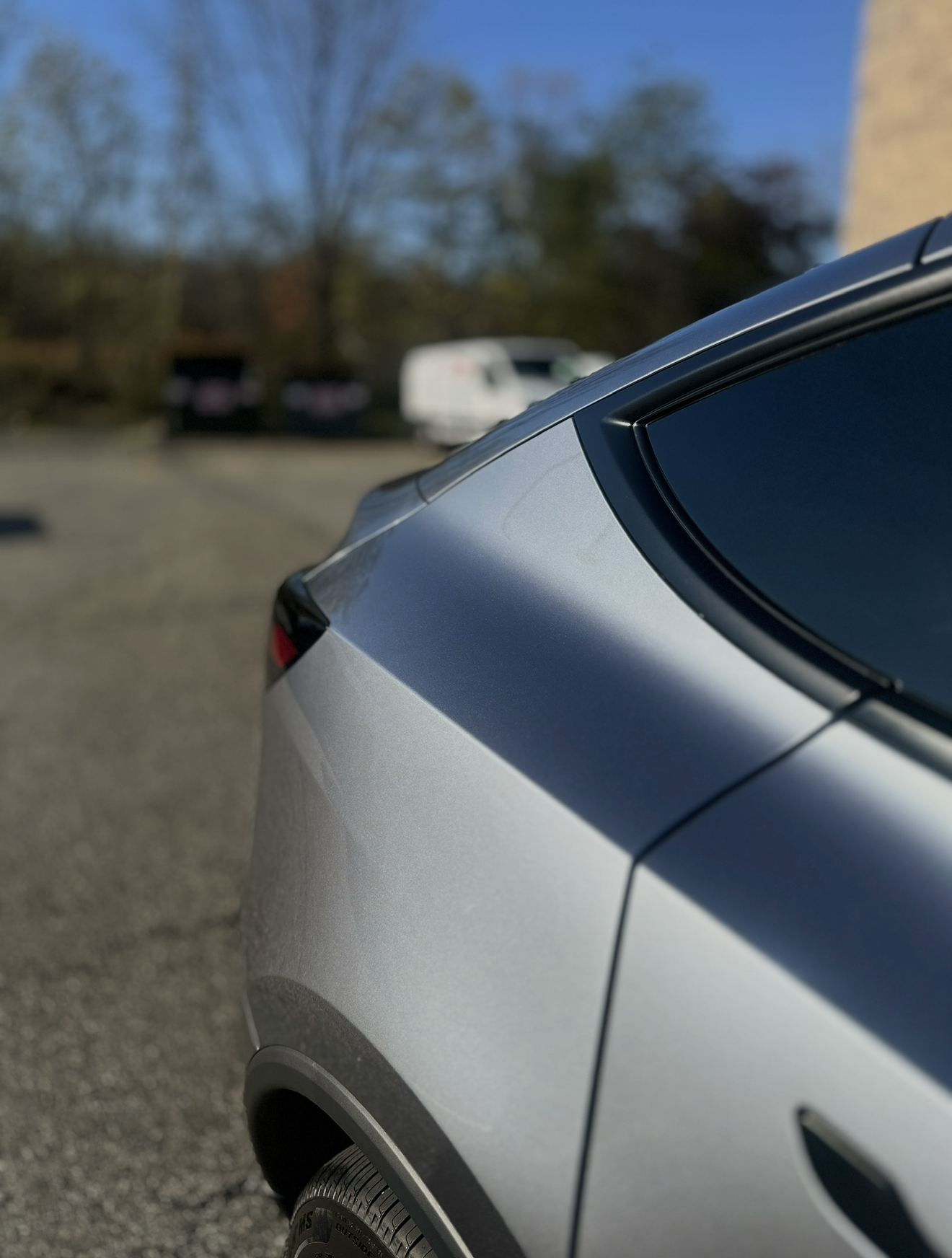 Close-up of a silver car’s rear side panel and window in a parking lot, with trees blurred in the background
