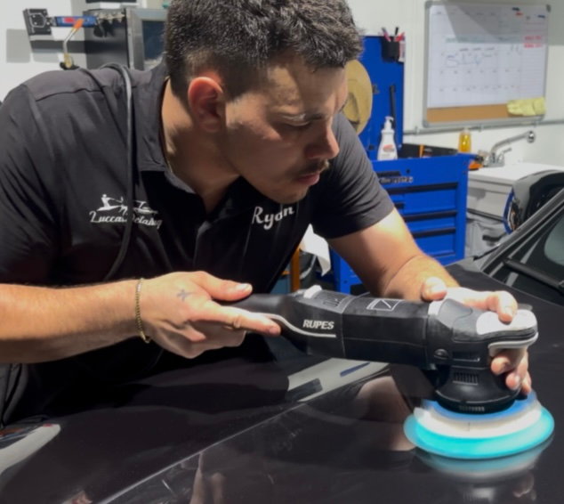 Detailer polishing a black car hood with a blue buffer in a workshop.