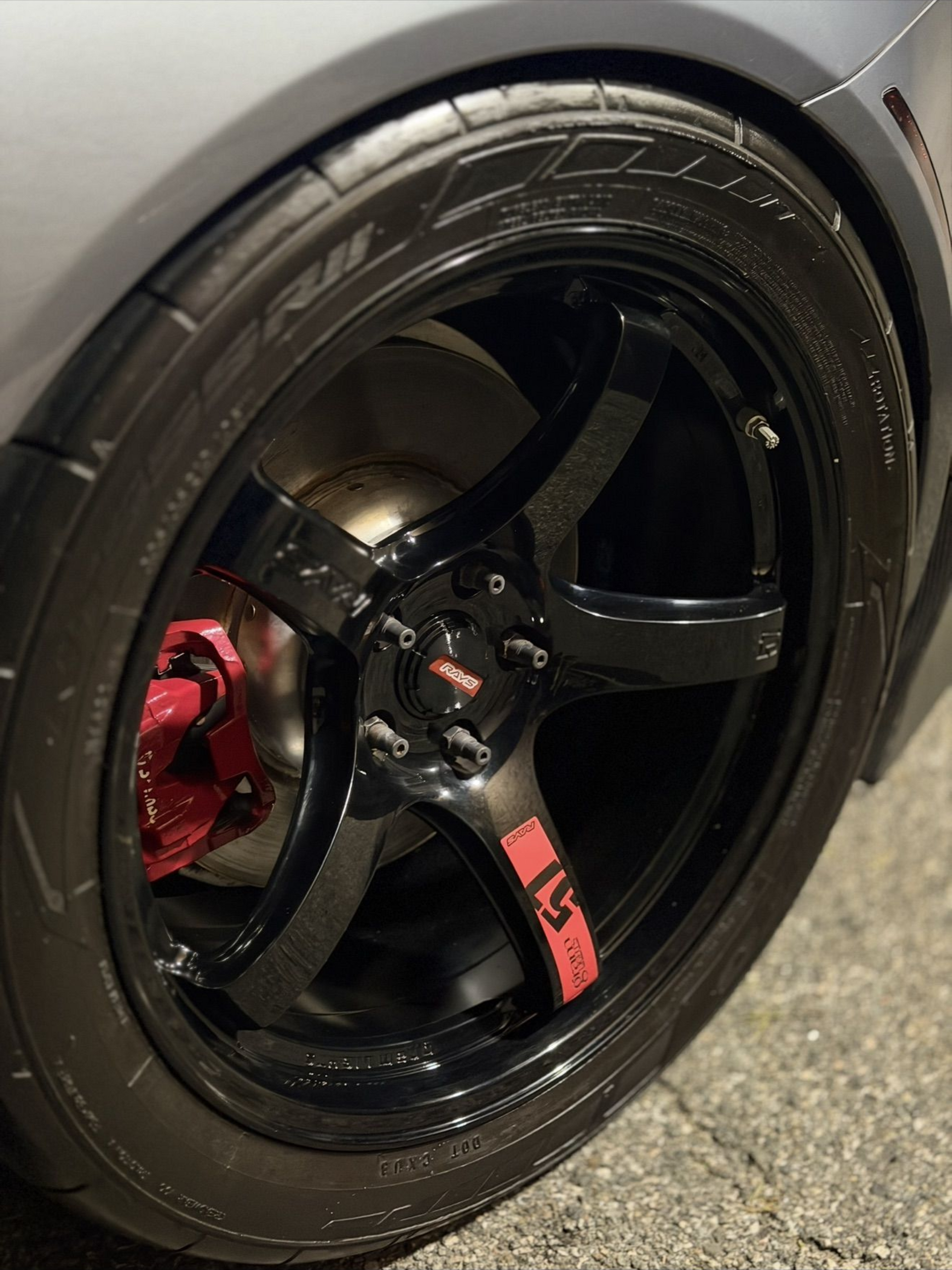 Close-up of a black car wheel with glossy rim and red brake caliper on pavement.