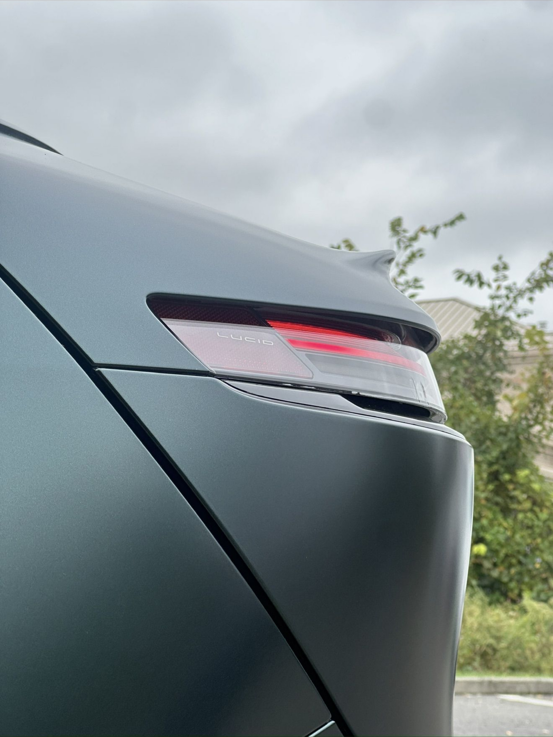 Close-up of a dark car’s rear taillight and bumper against a cloudy roadside background