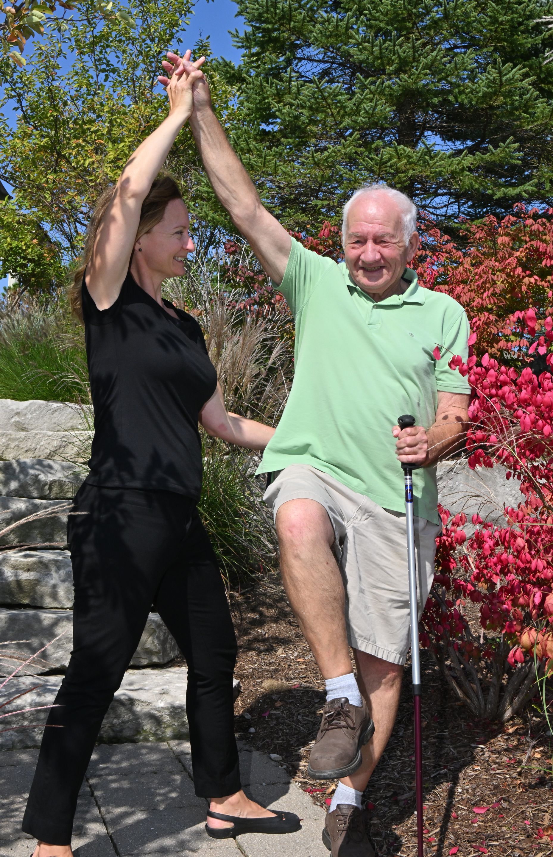 Physiotherapist supporting her elderly client as he lifts his right arm and knee to balance on his left leg. They are practicing in a park surrounded by beautiful foliage.