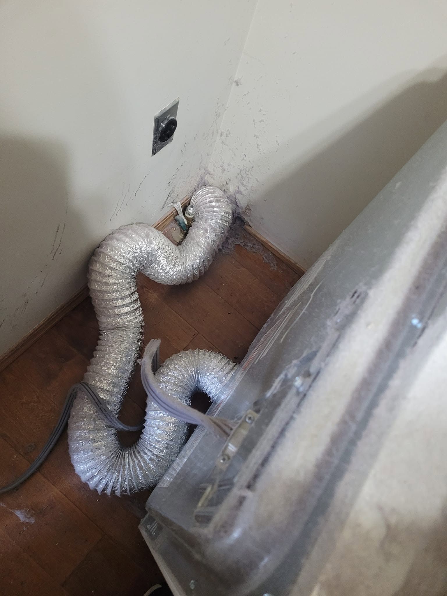 Silver ducting hose on a wooden floor beside a wall outlet and appliance panel
