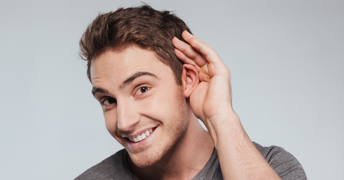 Smiling person with short brown hair cupping a hand to their ear against a gray background