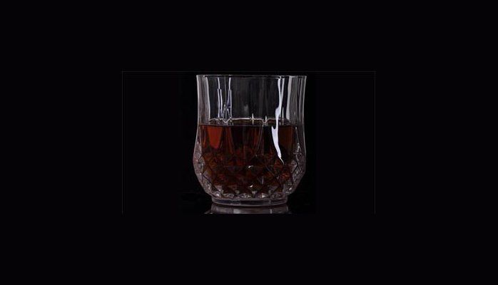 A clear glass tumbler with a dark amber drink against a black background.