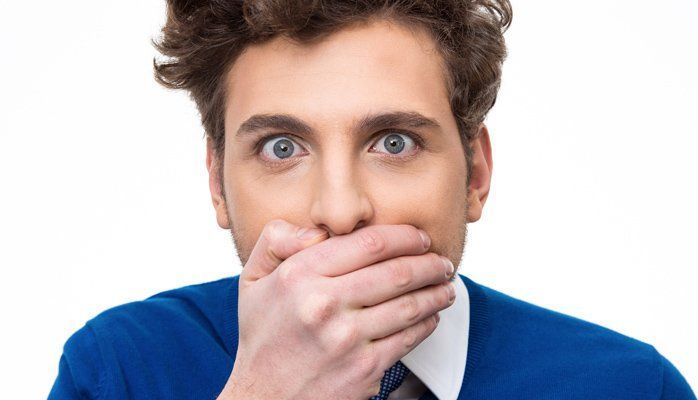 Man with blue shirt covering his mouth in surprise against a white background