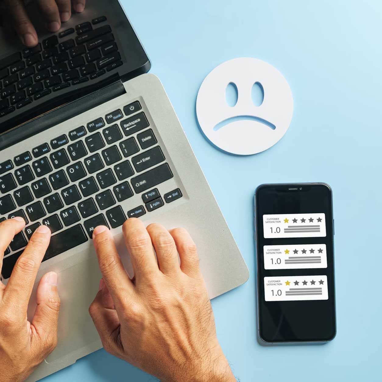 Hands typing on laptop beside smartphone with ratings and sad face icon on blue background